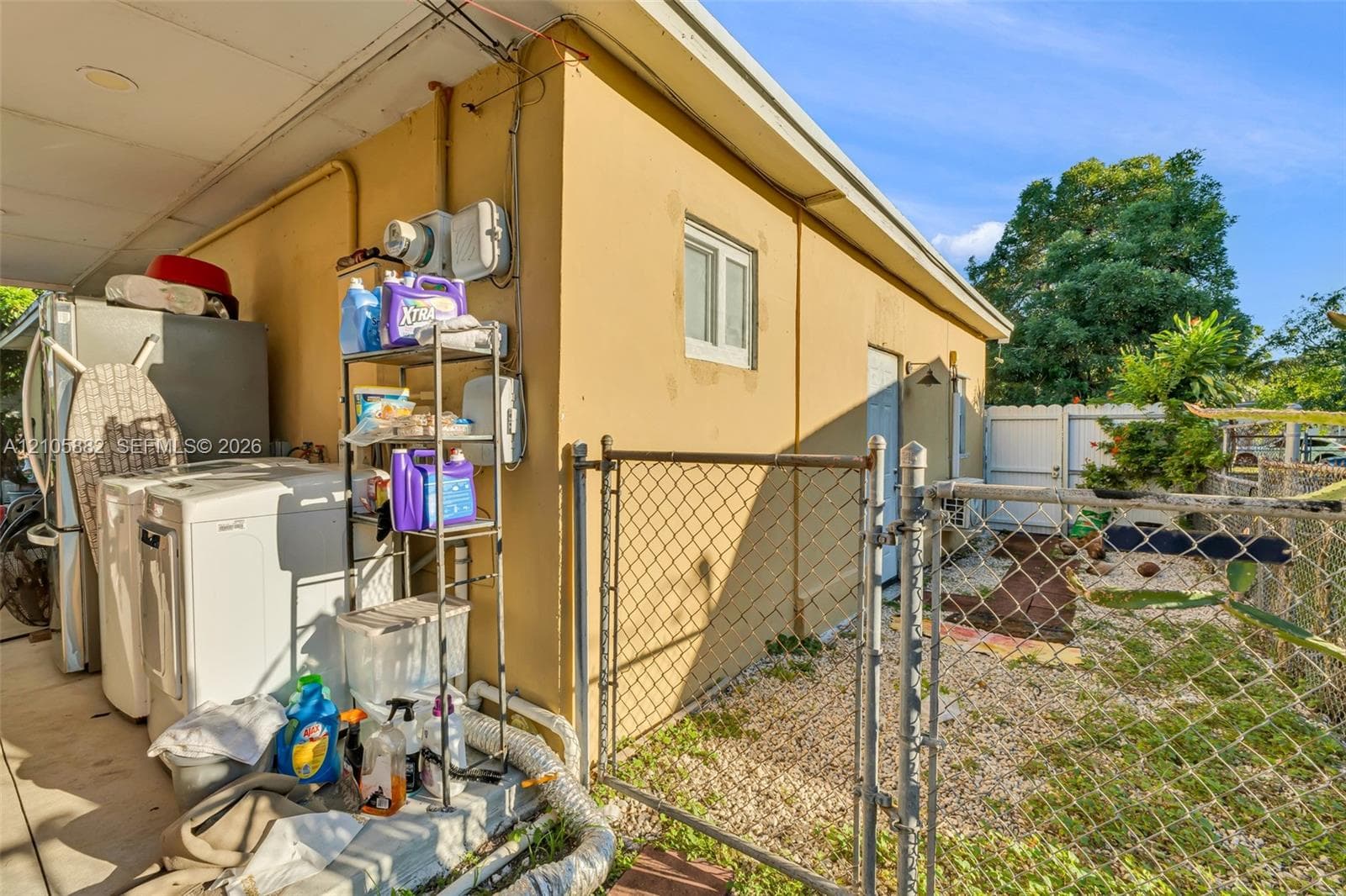 Exterior service area featuring a covered concrete patio, stucco siding, and a chain-link fenced perimeter with gravel ground cover