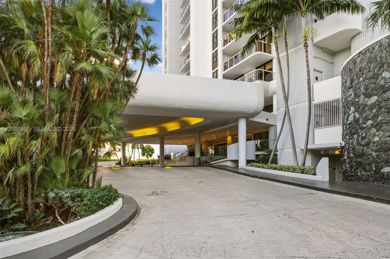 Covered porte-cochère entry featuring stone masonry accents, a concrete drive, and mature palm landscaping