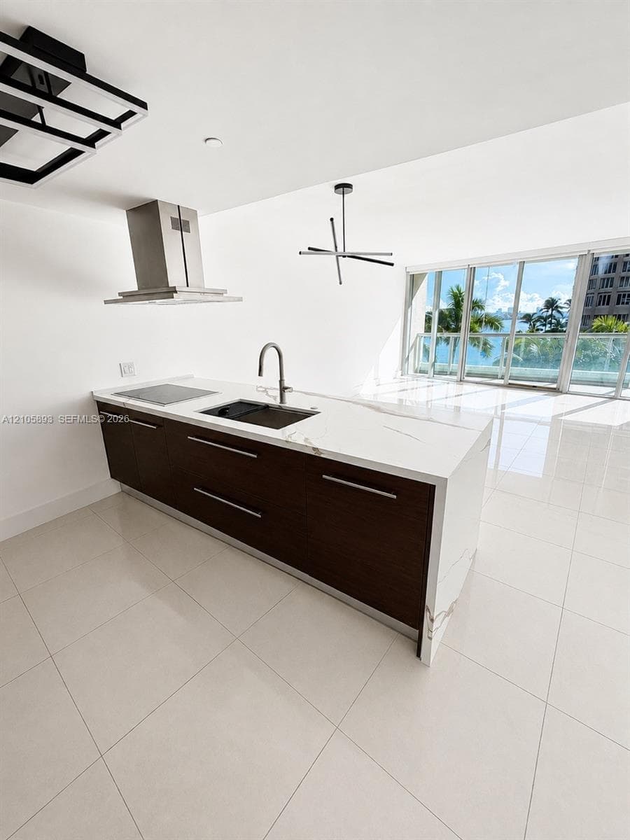 Kitchen island with white stone countertop, undermount sink, dark wood-finish cabinetry, stainless steel range hood, and large-format tile flooring