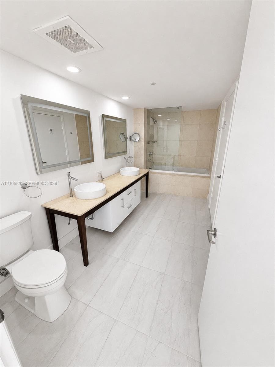 Dual-vanity featuring vessel sinks, stone-finish countertop, custom cabinetry, large-format tile flooring, and a glass-enclosed shower-tub combination