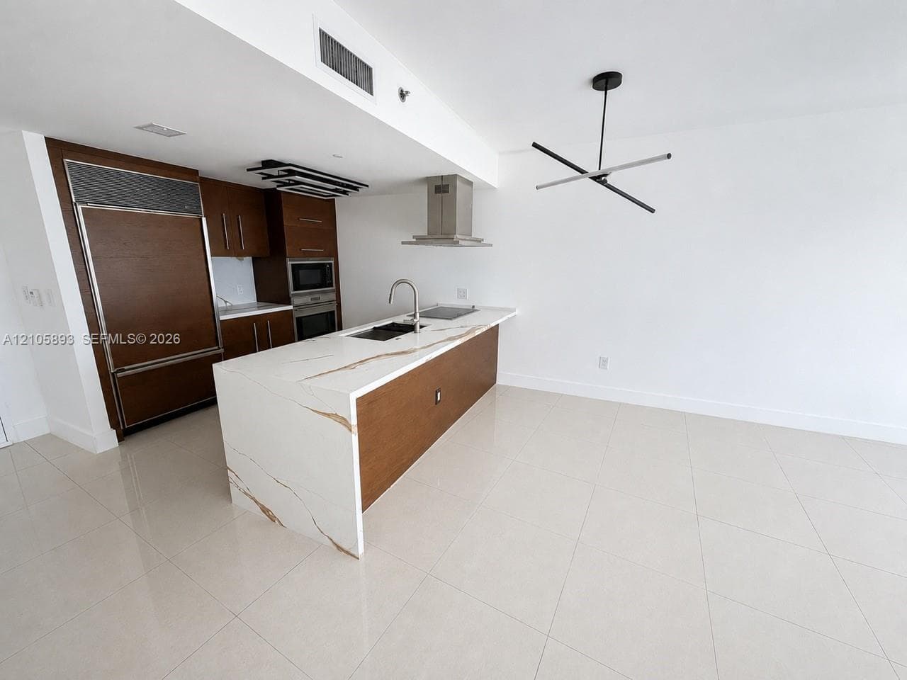 Modern kitchen featuring a waterfall island with stone-finish countertop, wood-finish cabinetry, stainless steel range hood, built-in panel-ready appliances, and a minimalist geometric chandelier