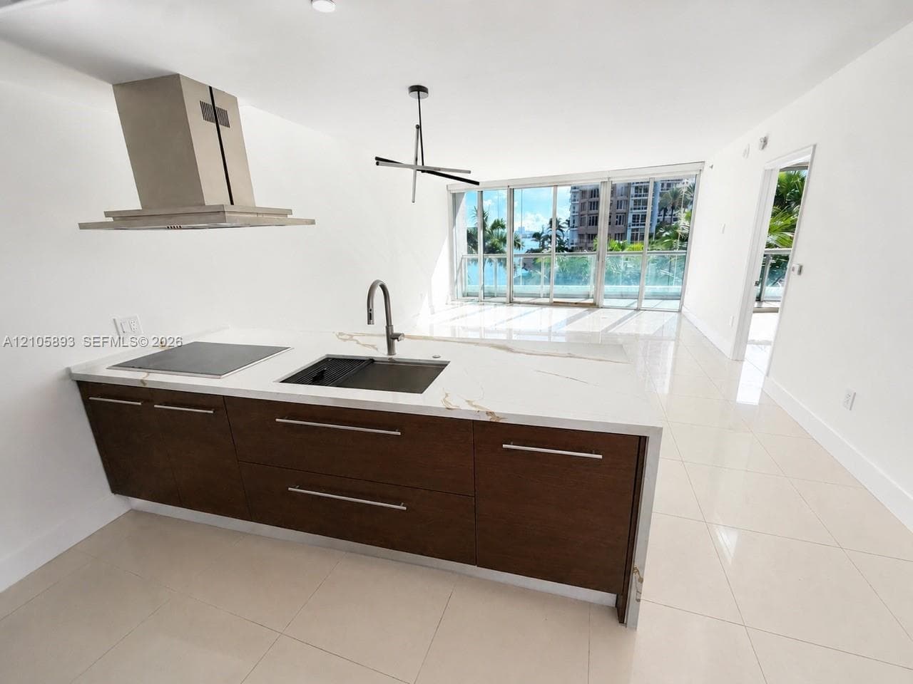 Kitchen island featuring a waterfall countertop with integrated cooktop and undermount sink, dark wood-finish cabinetry, stainless steel vent hood, and large-format tile flooring