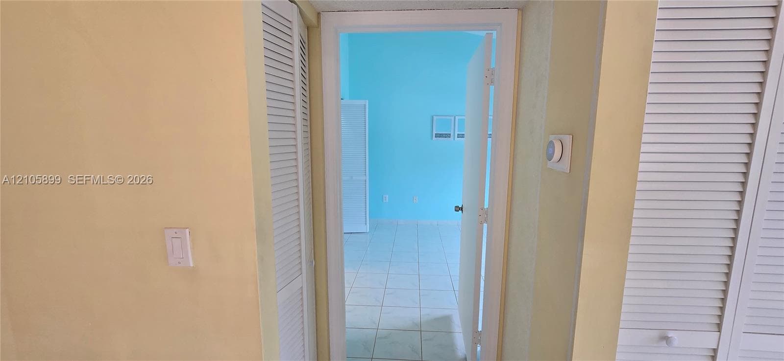 Interior doorway featuring white louvered closet doors, wall-mounted thermostat, and light-toned tile flooring