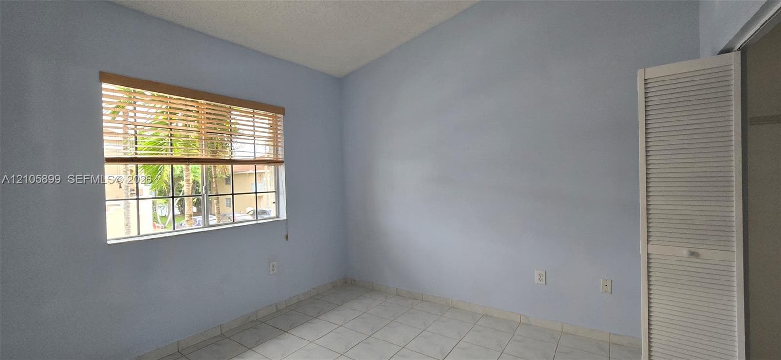 Room featuring neutral-toned ceramic tile flooring, a large window with slatted blinds, and a built-in louvered-panel closet door