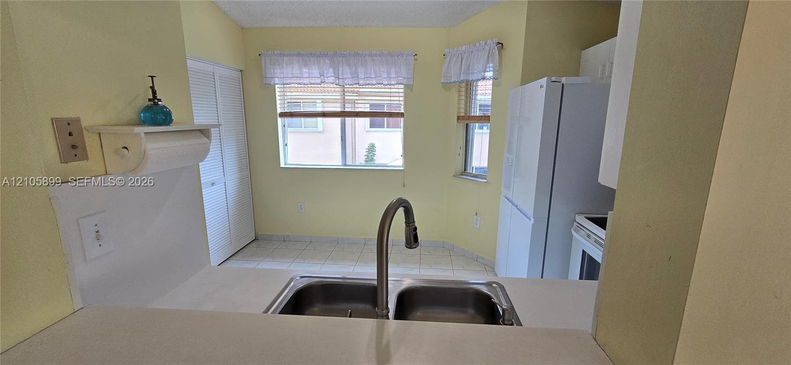 Kitchen area featuring a dual-basin stainless steel sink with a high-arch gooseneck faucet, tile flooring, white louvered closet doors, and windows with wood-slat blinds