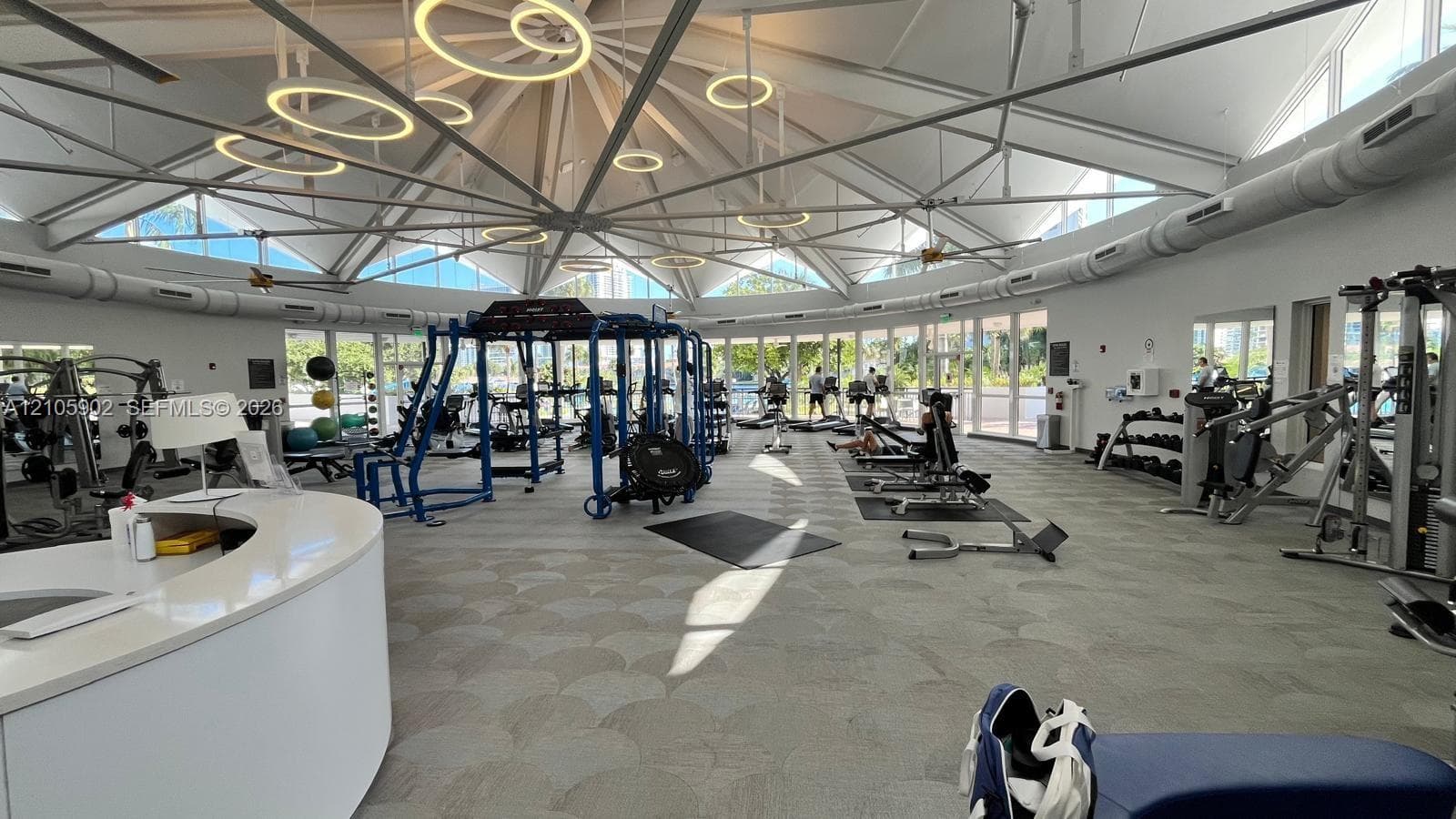 Expansive fitness center featuring a high-pitched vaulted ceiling, exposed structural steel trusses, circular pendant lighting, and a wrap-around wall of glass windows