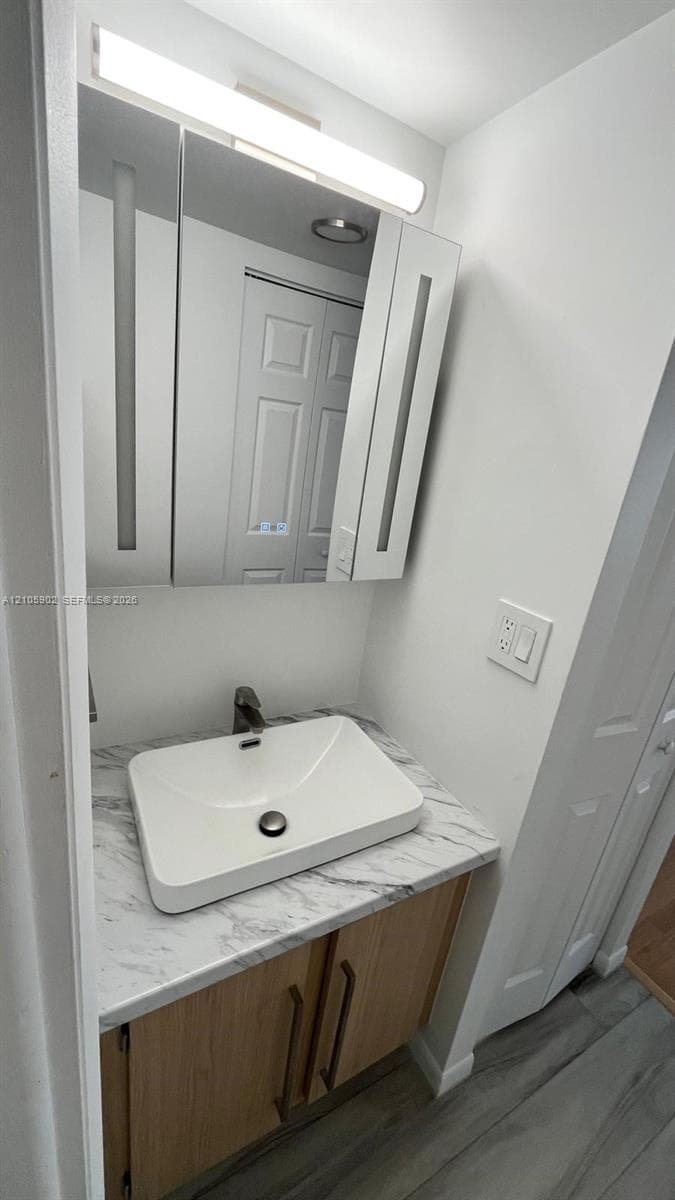 Vanity featuring a stone-finish countertop, rectangular vessel sink, wood-tone cabinetry with vertical hardware, and an integrated mirrored medicine cabinet with linear lighting