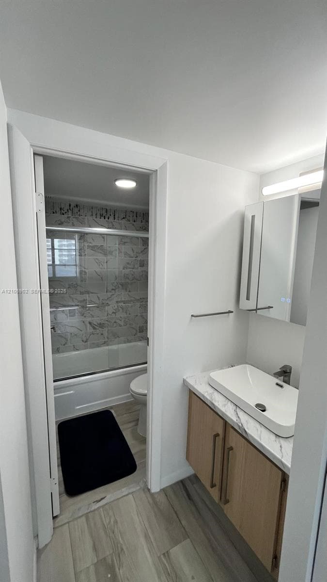 Vanity featuring a wood-finish cabinet, stone-patterned countertop, and rectangular vessel sink, complemented by wood-finish flooring and a tiled bathtub surround