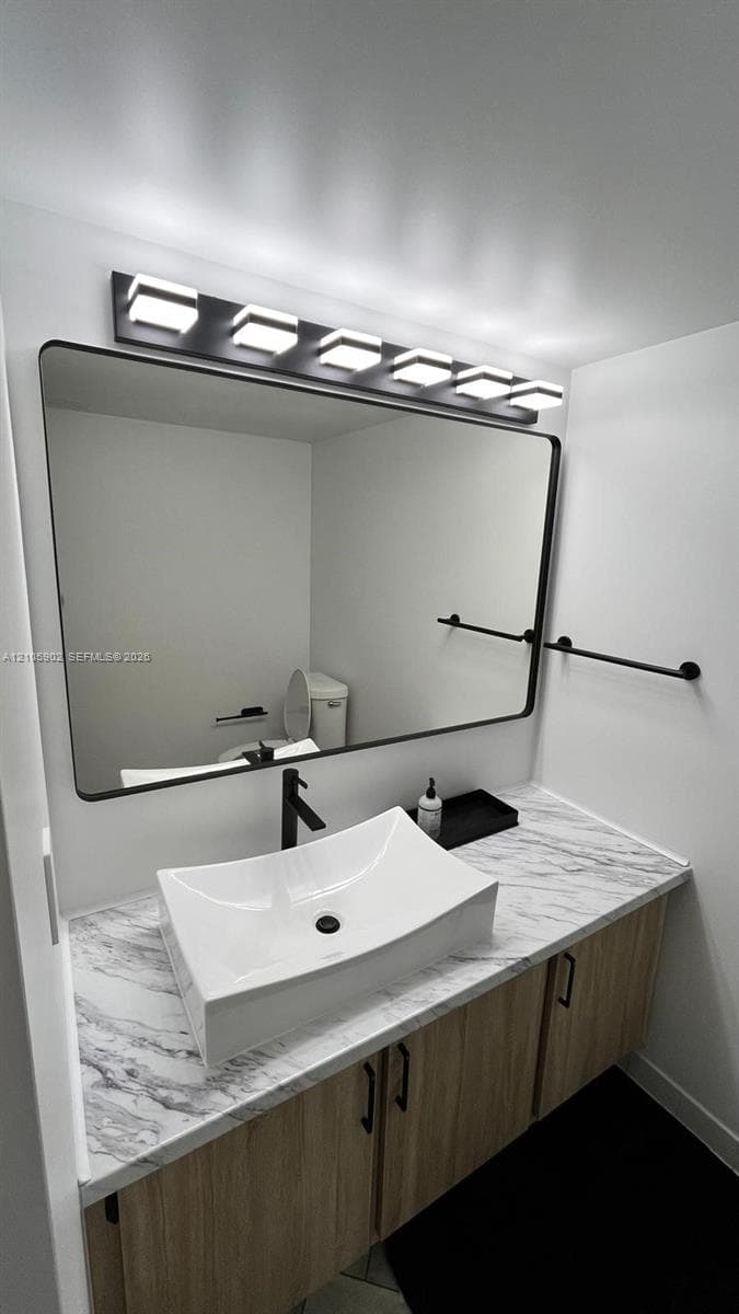 Floating vanity with wood-finish cabinetry, stone-patterned countertop, rectangular vessel sink, matte black faucet, and modern multi-light fixture