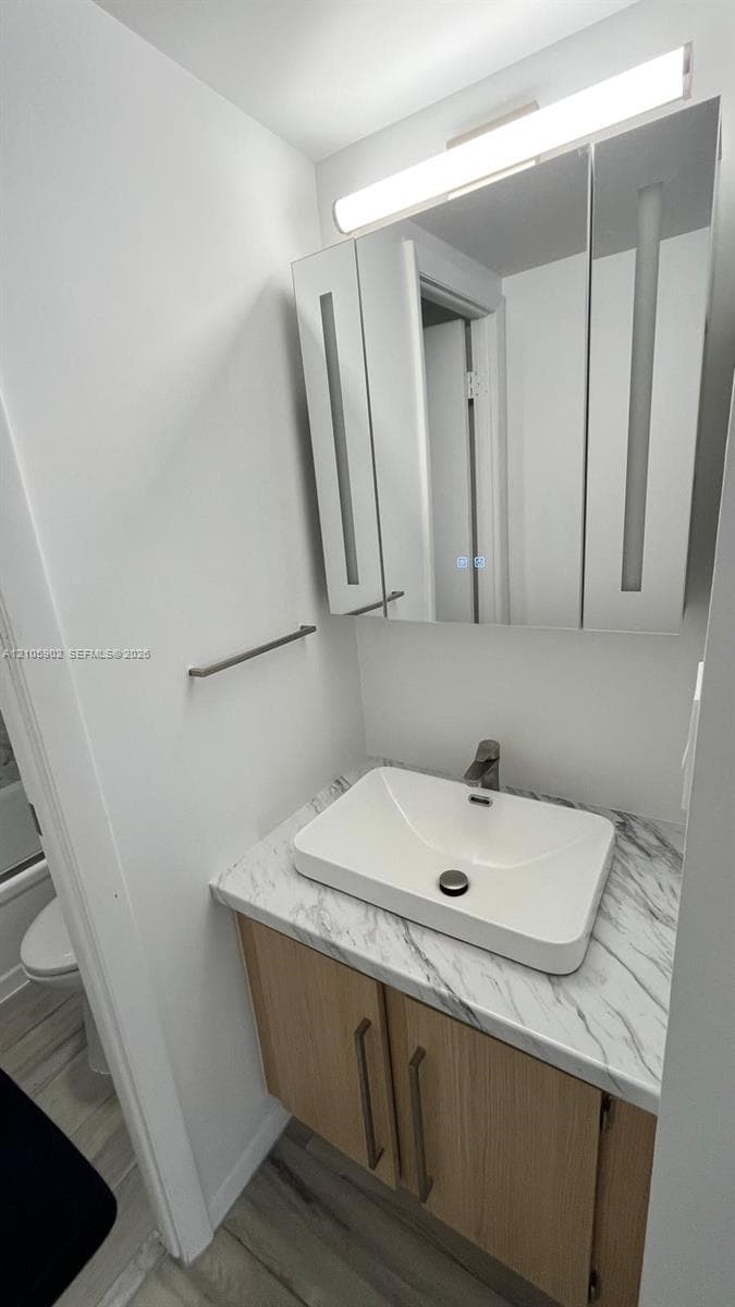 Vanity featuring a wood-finish cabinet, stone-patterned countertop, vessel sink, integrated medicine cabinet with mirrored doors, and an overhead light fixture