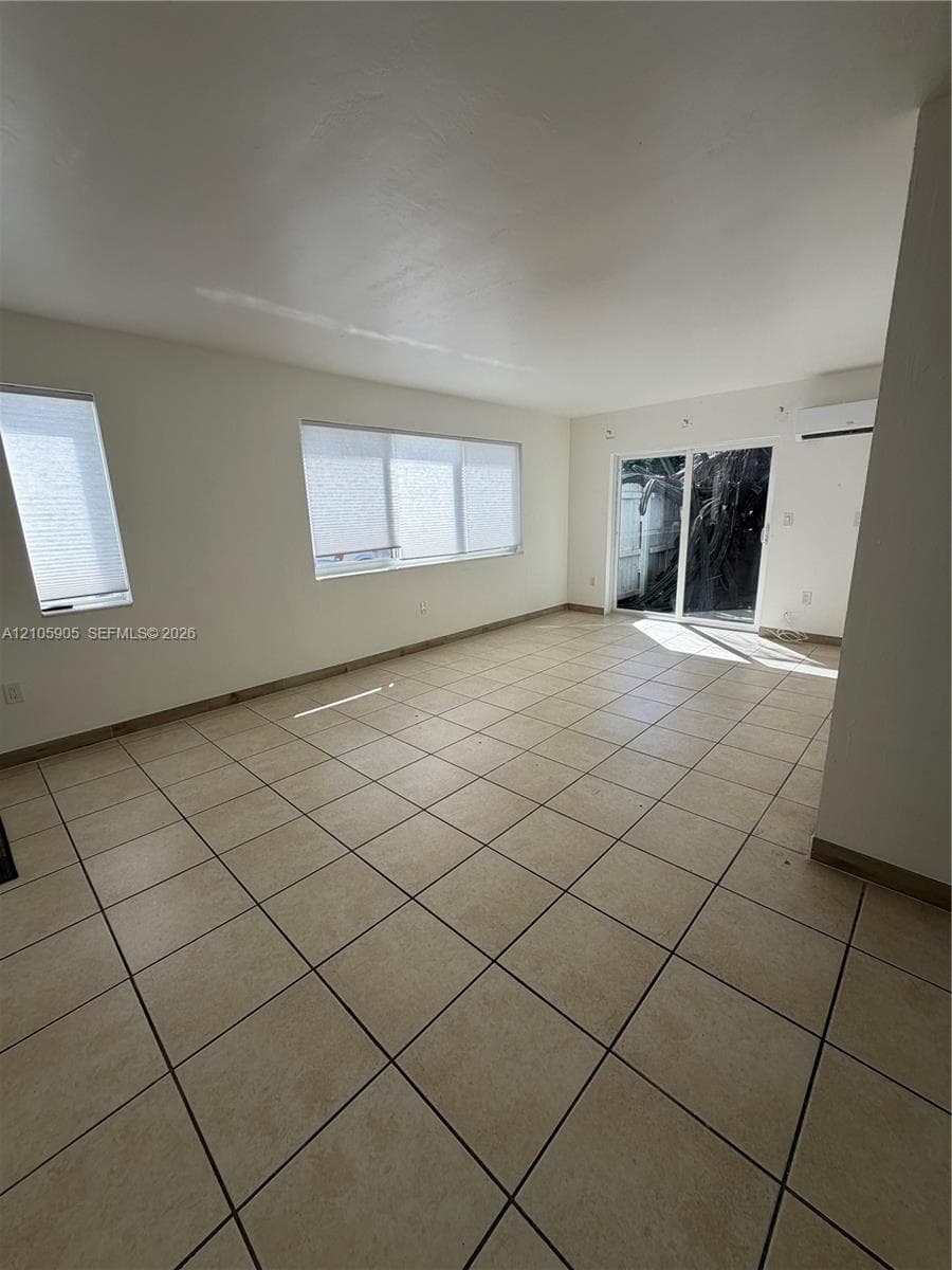 Neutral-toned tile flooring, large multi-pane windows, sliding glass door, and wall-mounted climate control unit