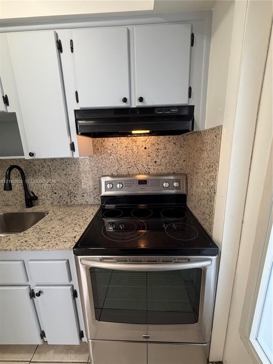 Kitchen featuring light-toned cabinetry, stone-finish countertops, a stainless steel electric range, a black range hood, and a dark-finish faucet