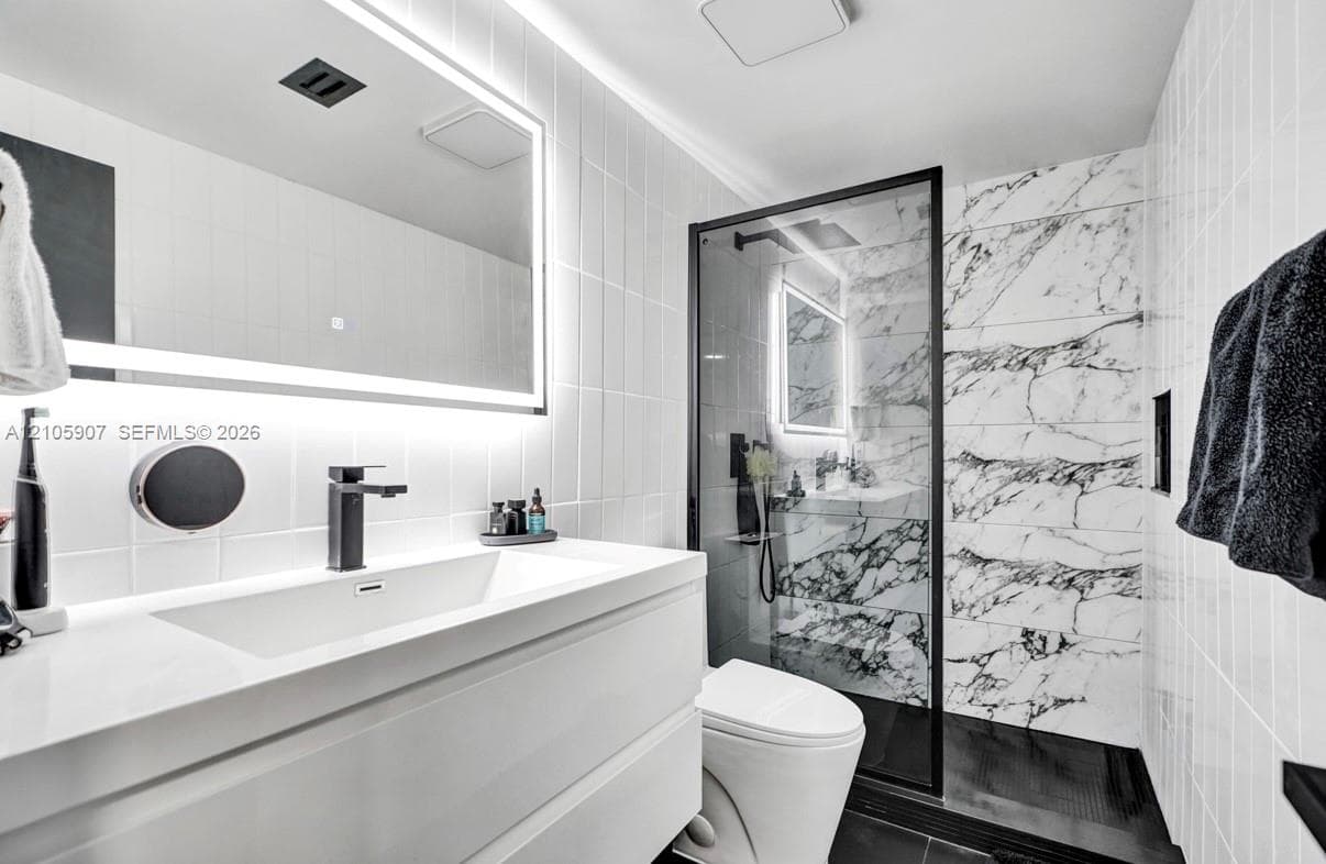 Modern bathroom featuring a floating vanity with an integrated rectangular sink, wall-mounted matte black faucet, illuminated vanity mirror, and a walk-in shower with stone-patterned wall tile and black metal framing