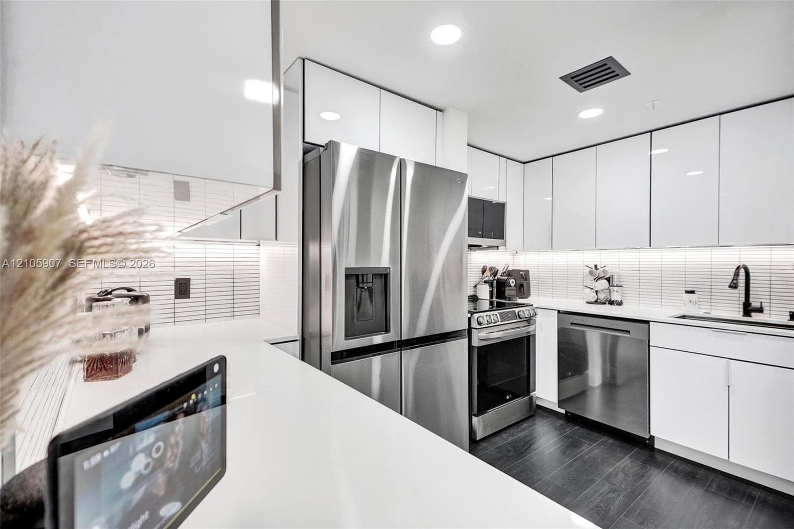 Kitchen featuring high-gloss white cabinetry, stainless steel appliances, white solid-surface countertops, linear tile backsplash, and dark wood-finish flooring