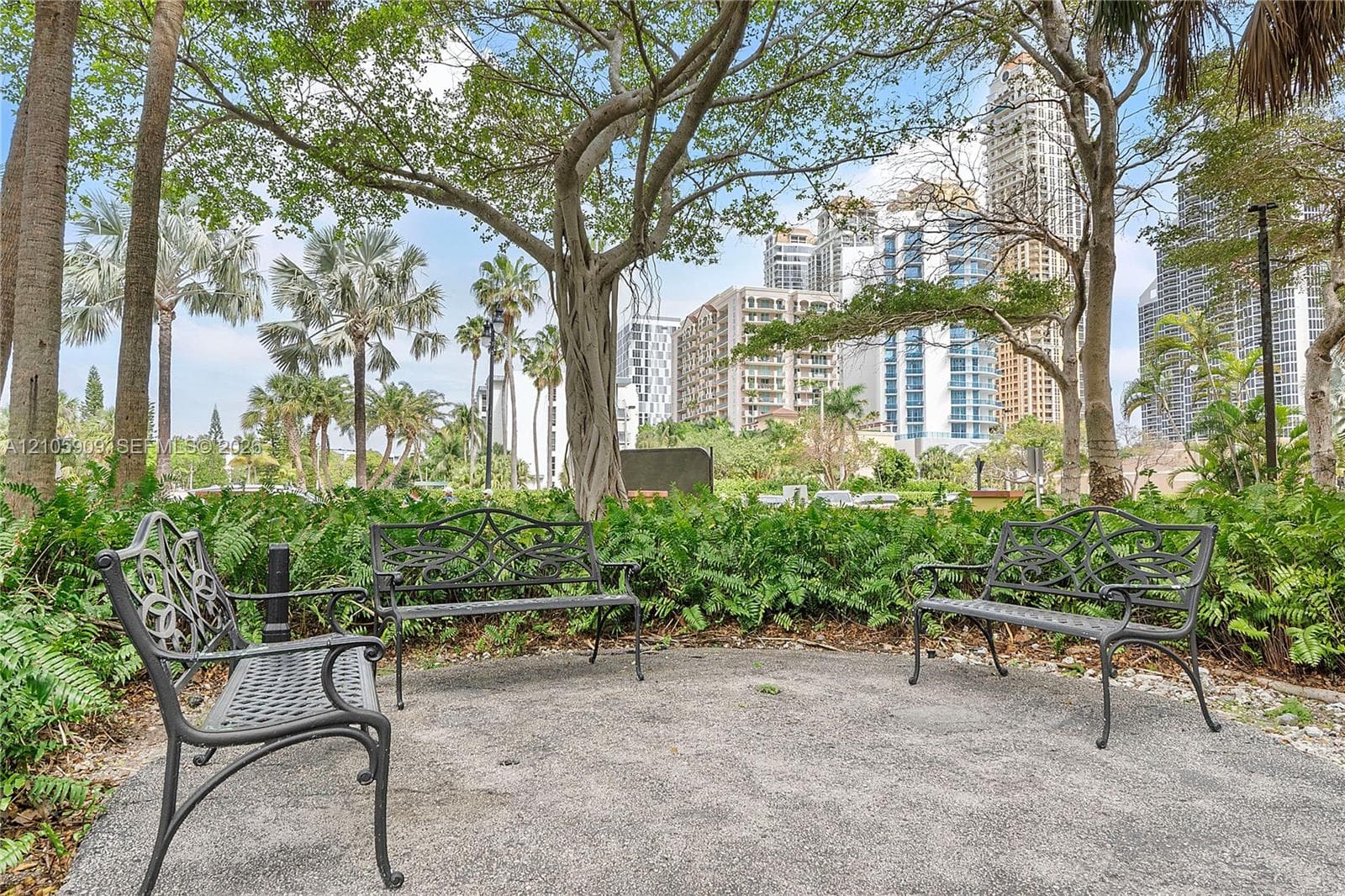 Landscaped grounds featuring crushed stone pathways, mature native trees, tropical foliage, and decorative wrought-iron benches