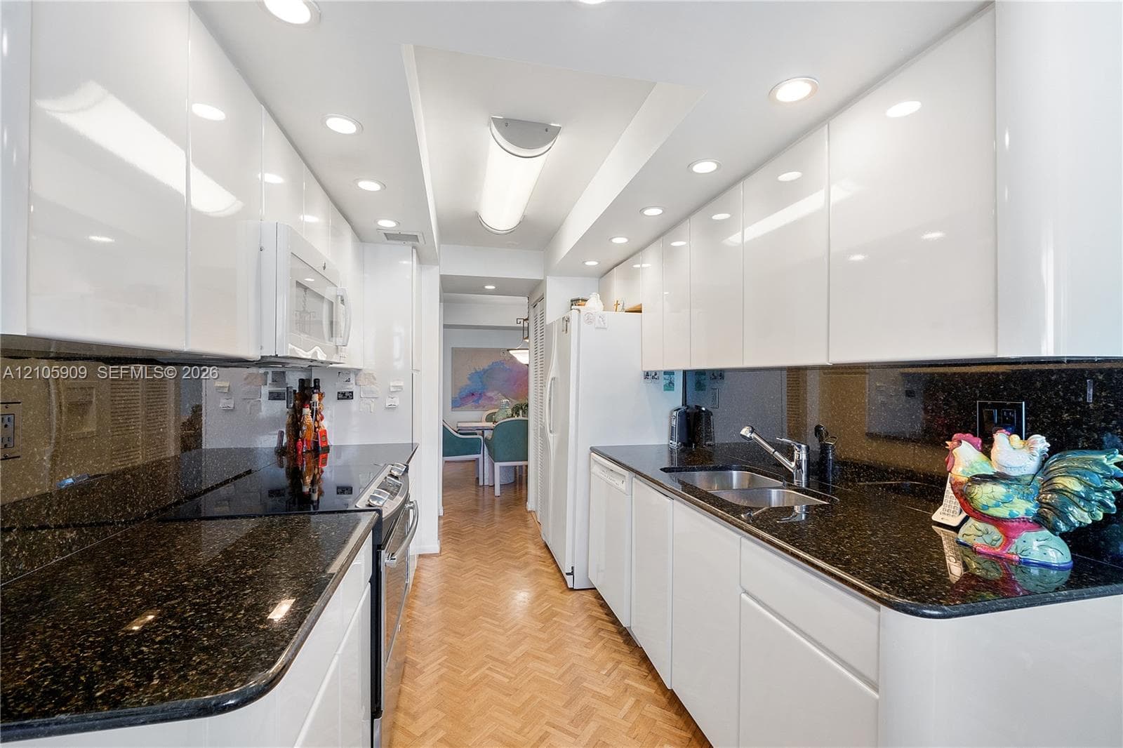 Kitchen featuring high-gloss white cabinetry, dark stone countertops, stainless steel dual-basin sink, built-in microwave, and herringbone-patterned wood flooring