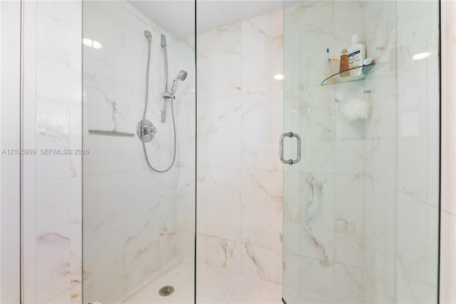 Walk-in shower featuring large-format stone-finish tile surround, frameless glass enclosure, adjustable hand-held showerhead, and integrated glass corner shelving