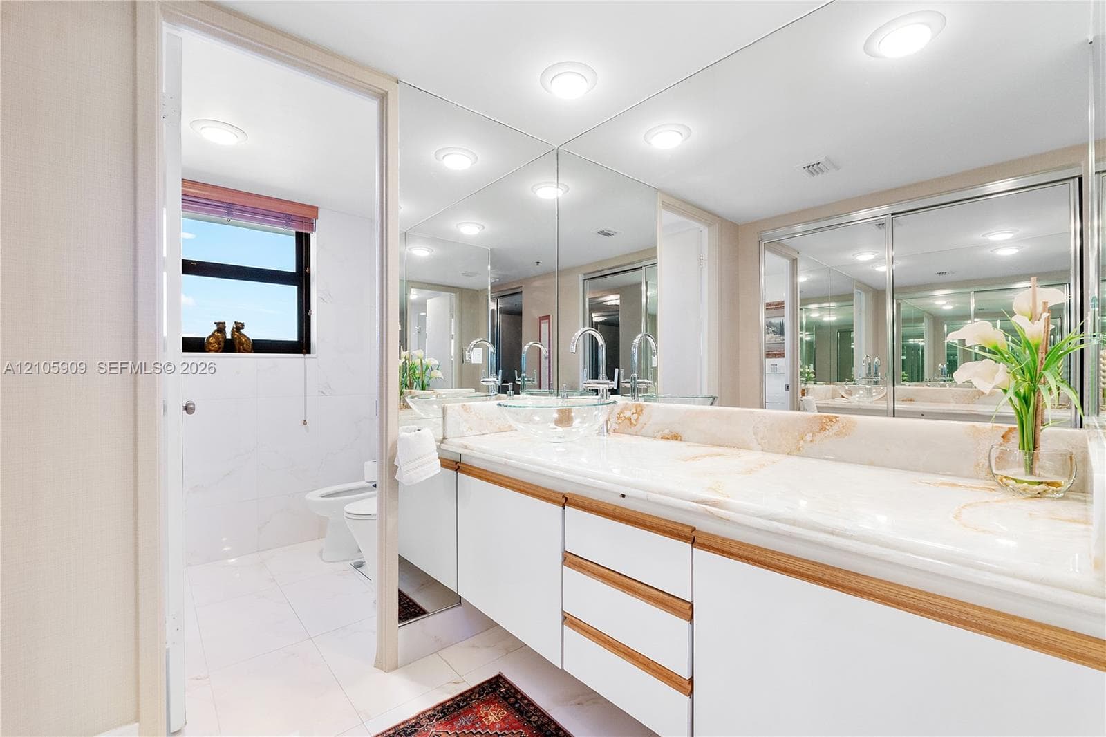 Full-width vanity featuring white cabinetry with wood-tone accents, stone-surface countertop, recessed ceiling lighting, and expansive wall-to-wall mirror