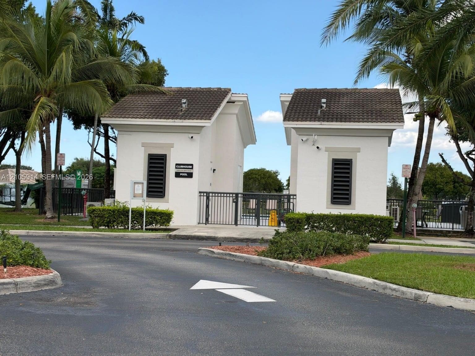 Gated entry featuring dual stucco structures, tile roofing, and metal security fencing surrounded by manicured landscaping and palm trees