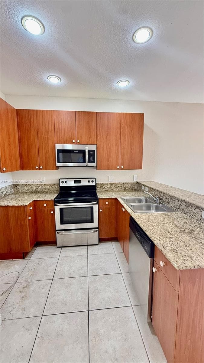 Kitchen featuring wood-finish cabinetry, stone-style countertops, stainless steel appliances, a dual-basin sink, and tile flooring