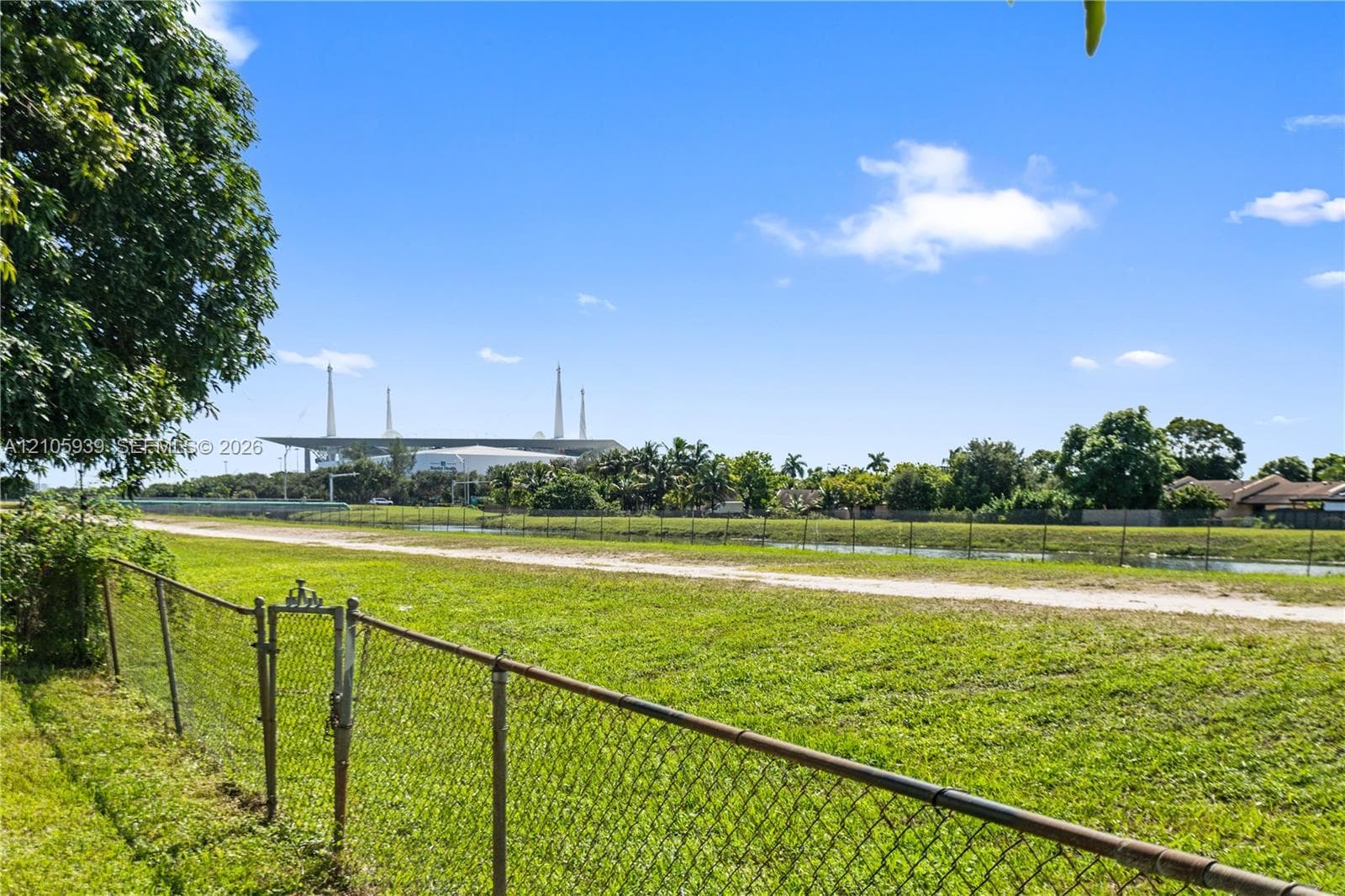 Expansive grassy lot enclosed by a chain-link fence, featuring open terrain, mature perimeter trees, and a waterway border