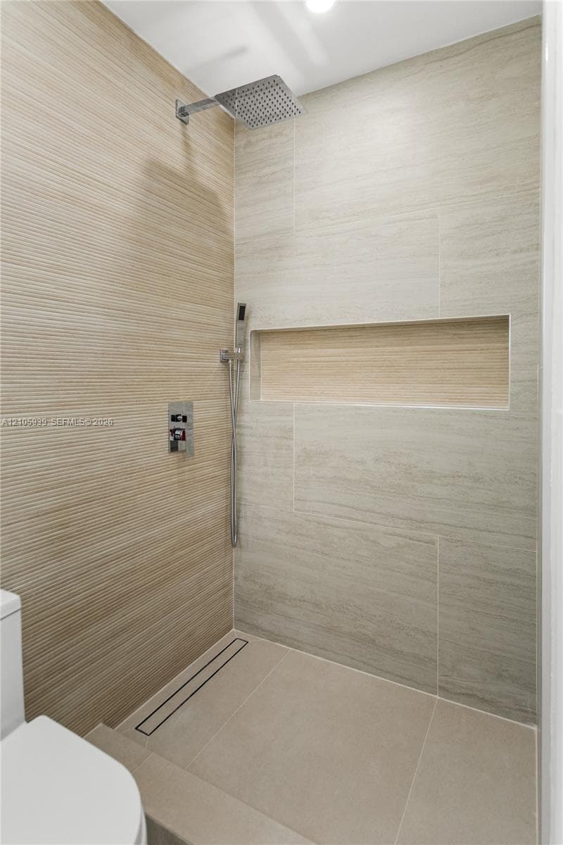 Walk-in shower featuring textured wall tile, large-format smooth wall tile, integrated recessed shelving, square rainfall showerhead with handheld sprayer, and a linear floor drain