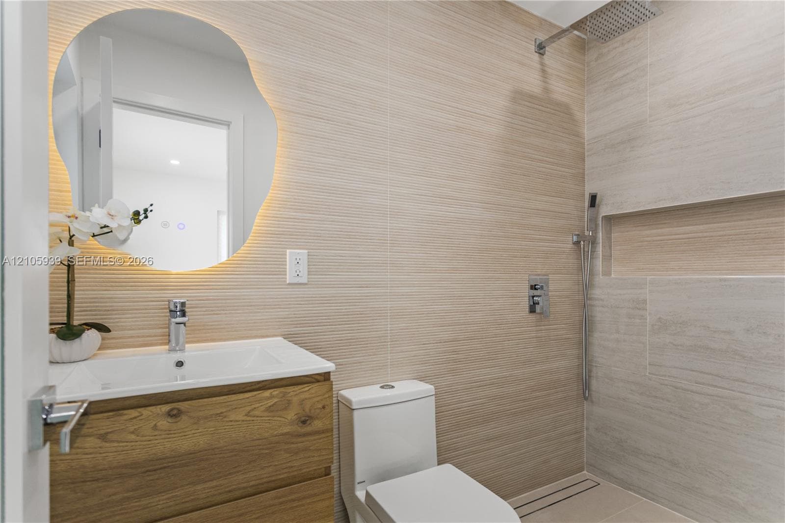 Wall-mounted wood-finish floating vanity, backlit organic-shaped mirror, horizontal textured wall tile, and walk-in shower featuring a built-in recessed niche and rainfall showerhead
