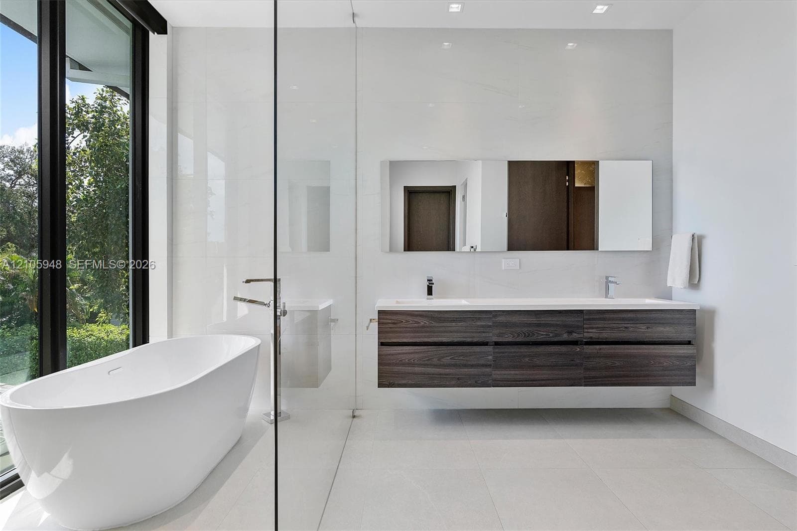 Freestanding oval soaking tub, wall-mounted dark wood-finish dual vanity with integrated sinks, large-format tile flooring, floor-to-ceiling glass-enclosed shower, and expansive window wall