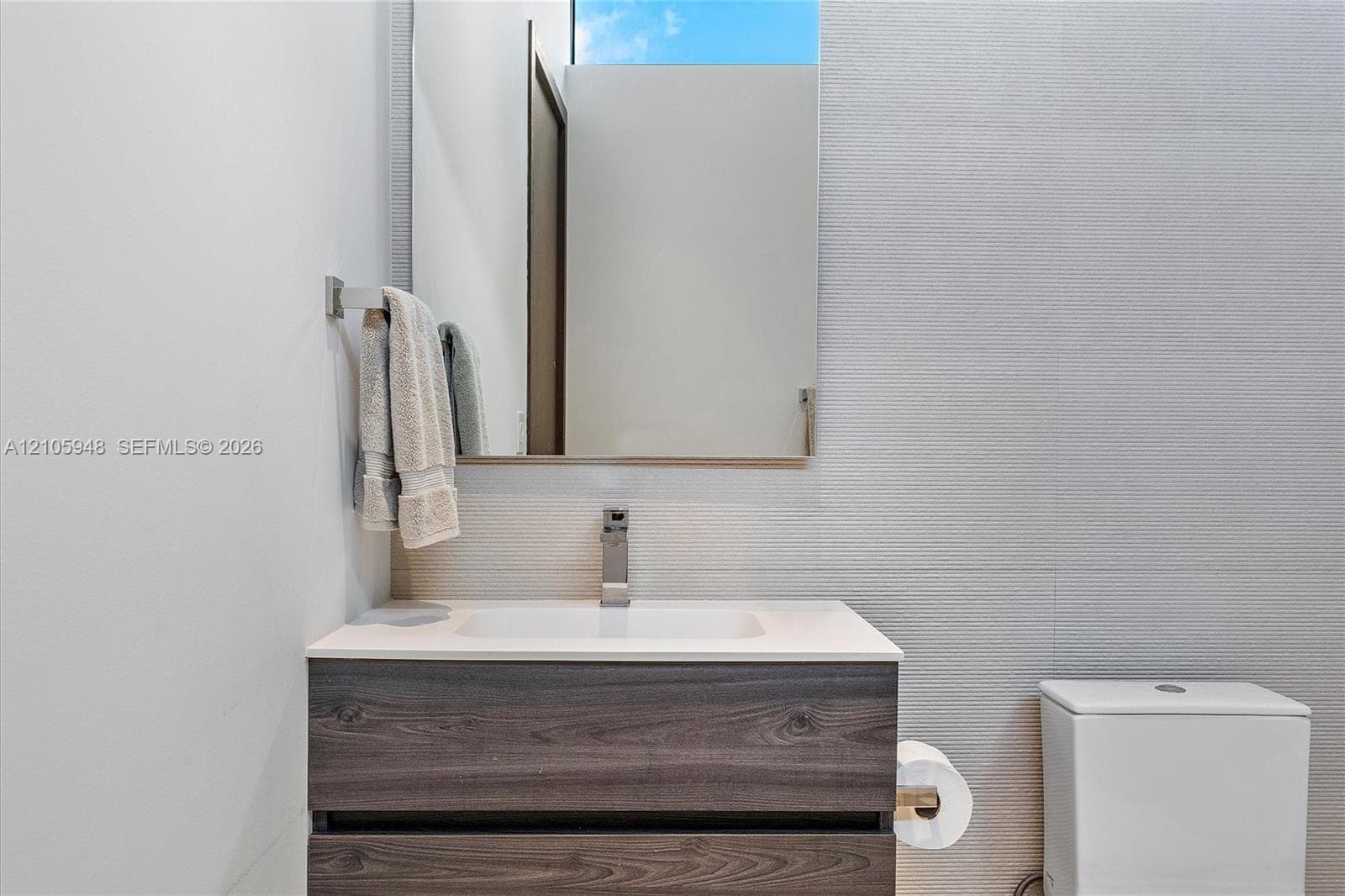 Floating vanity with a wood-finish, dual-drawer cabinet, crisp white countertop, integrated sink, and modern polished metal faucet, complemented by a large frameless mirror and textured wall tiling