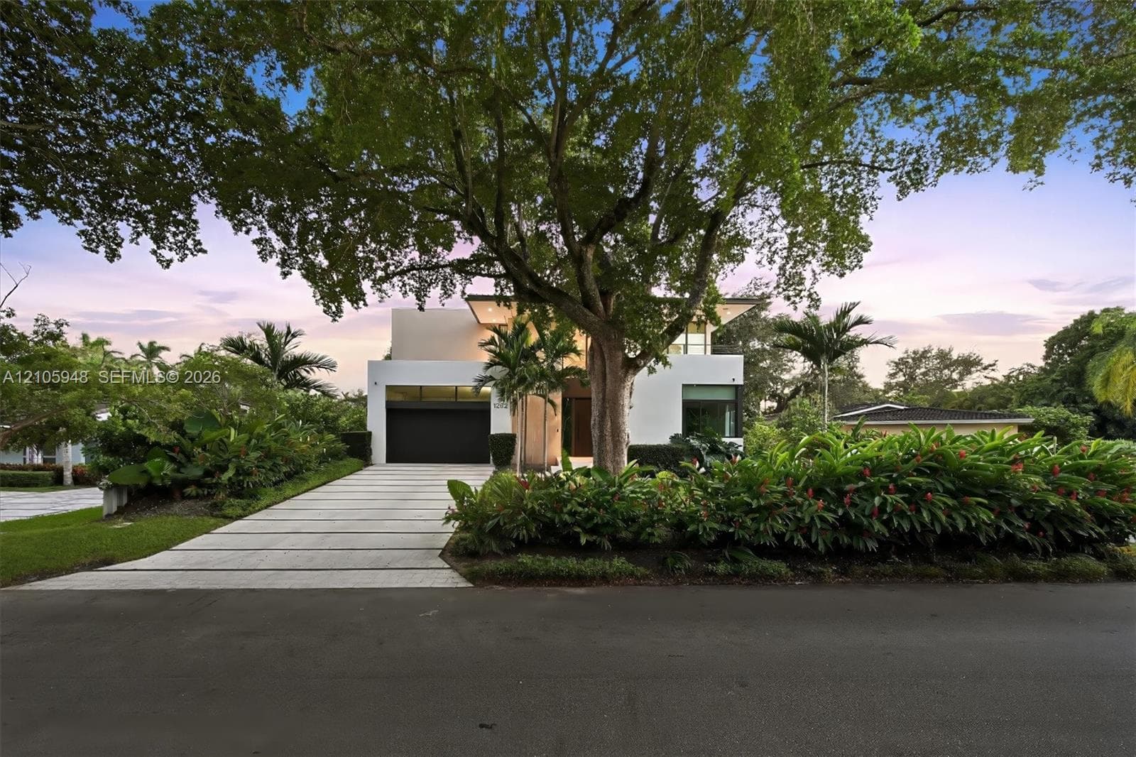 Modern architectural facade featuring a smooth stucco exterior, linear concrete paver driveway, dark-finish multi-car garage door, and lush tropical landscaping with mature canopy trees