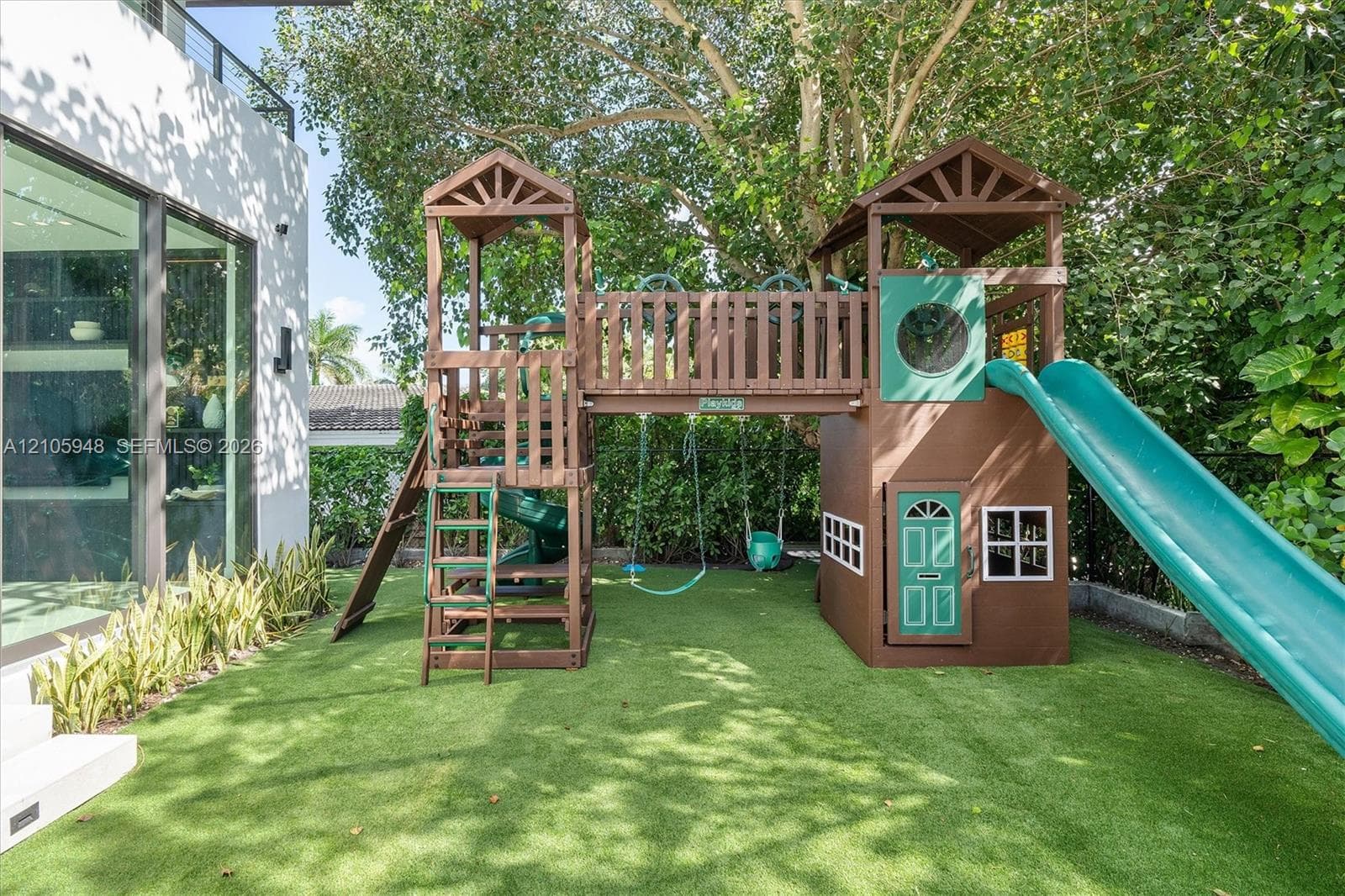 Outdoor area featuring low-maintenance synthetic turf, integrated wood play structure with dual towers and slide, and established perimeter privacy hedging
