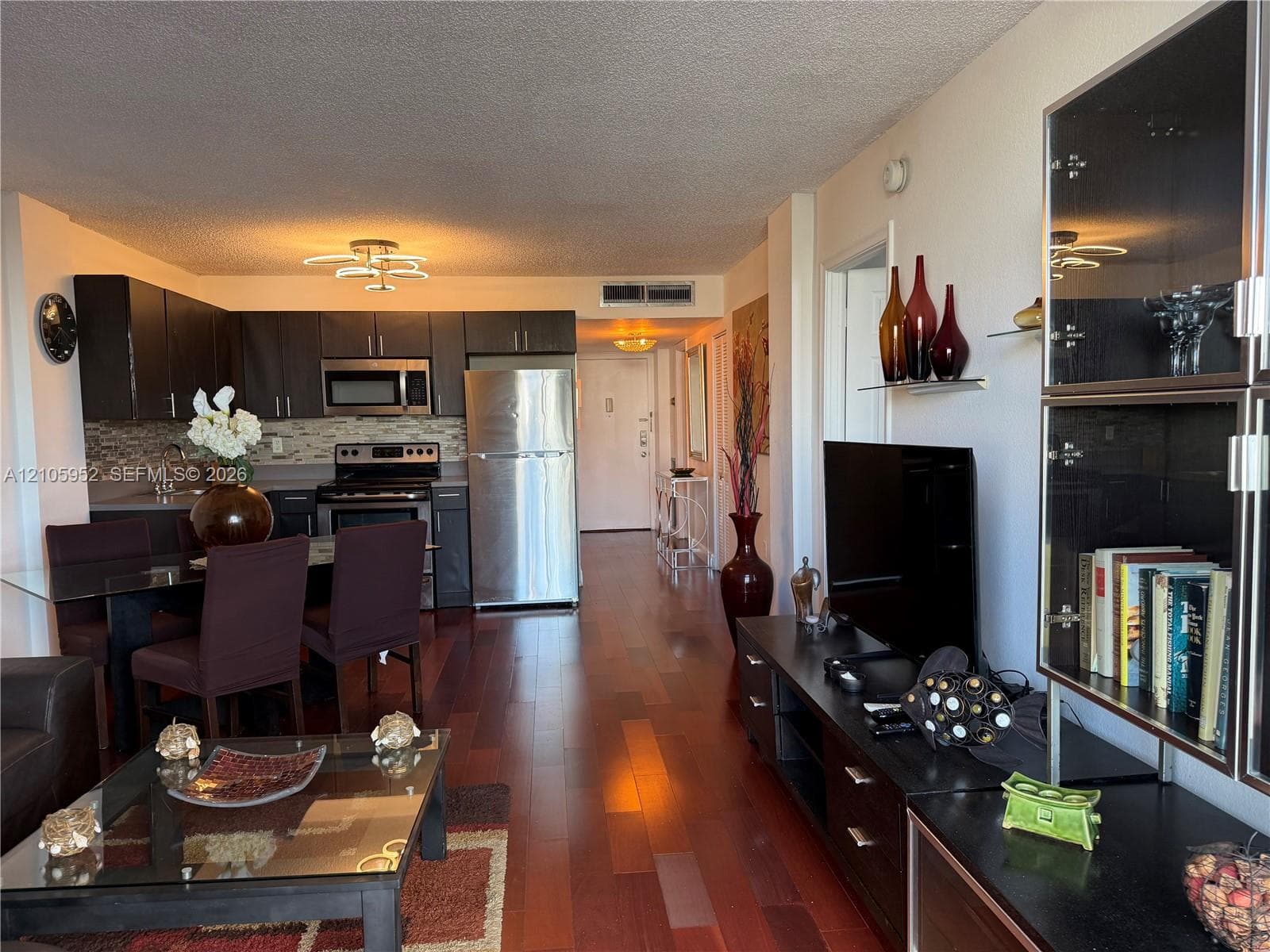 Open-concept living space featuring wood-finish flooring, dark cabinetry, stainless steel kitchen appliances, and decorative tile backsplash