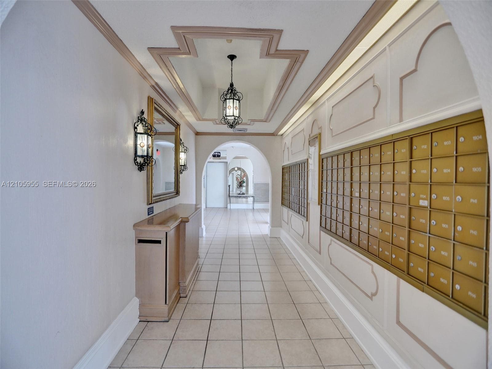 Entryway featuring ceramic tile flooring, built-in metal mailboxes, decorative wall molding, wrought-iron wall sconces, and a recessed ceiling design with a pendant fixture