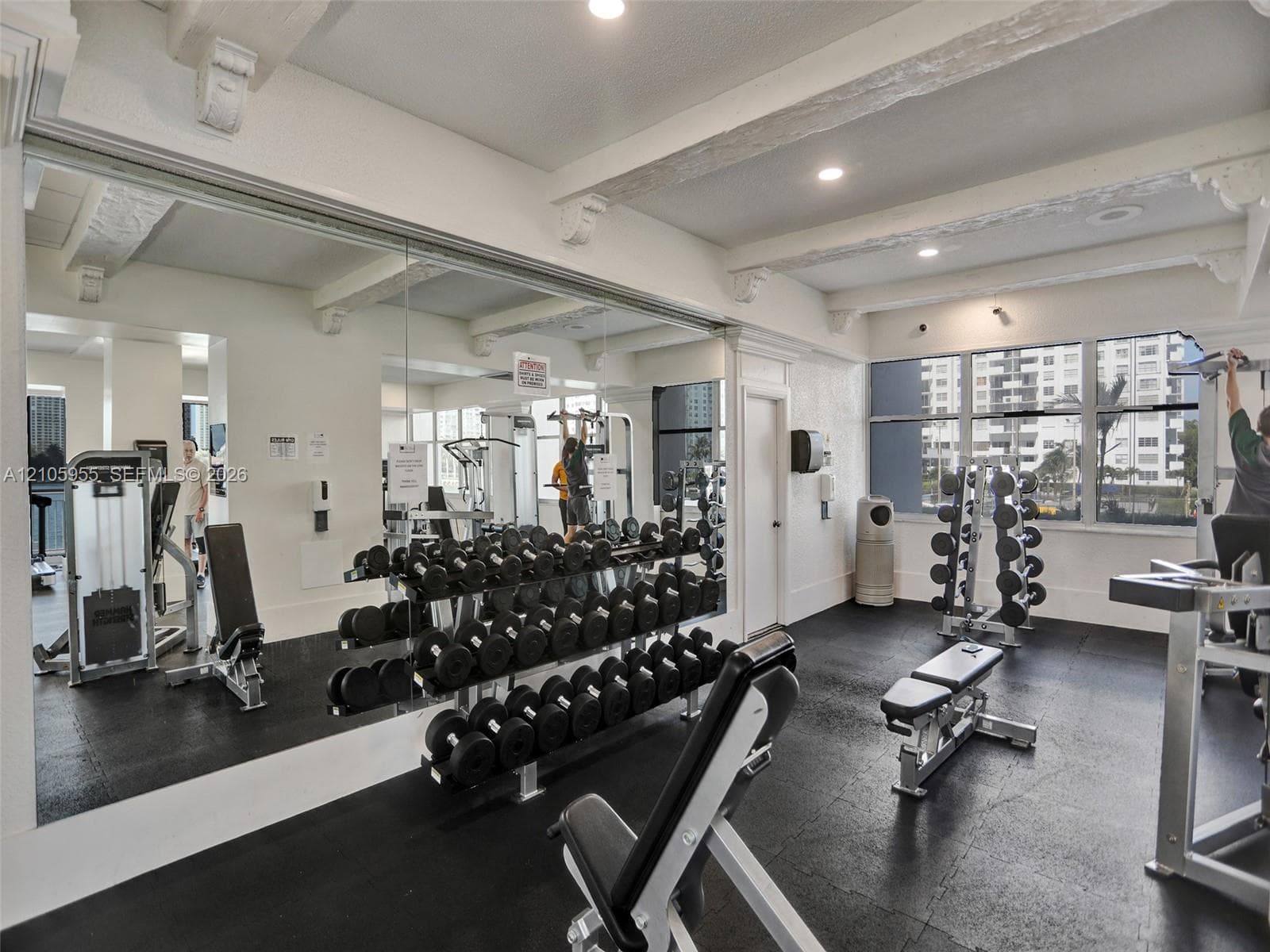 Fitness area featuring rubberized flooring, expansive wall-to-wall mirrors, exposed ceiling beams with corbel detailing, and oversized windows