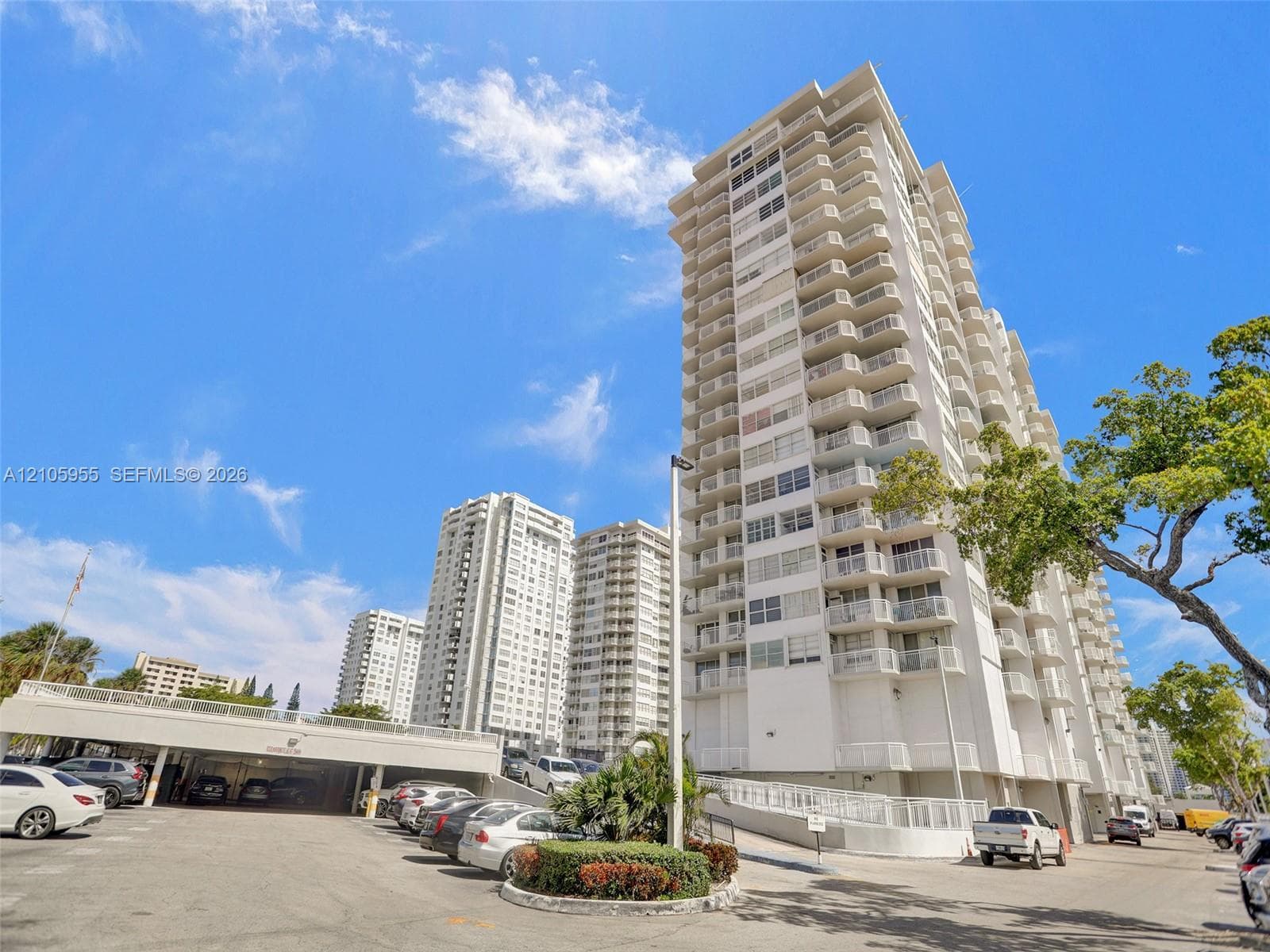 High-rise residential exterior featuring stacked private balconies, concrete facade, and integrated covered parking structure with landscaping