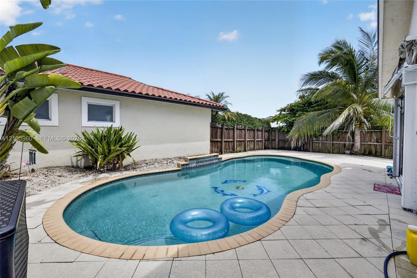 In-ground swimming pool featuring decorative tile accents and a stone-tiled patio, enclosed by a privacy fence and complemented by mature tropical landscaping