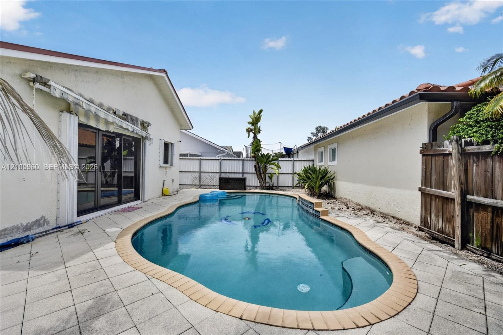 In-ground swimming pool with a tan coping surround, set within a tiled patio area enclosed by wood fencing and a white privacy fence