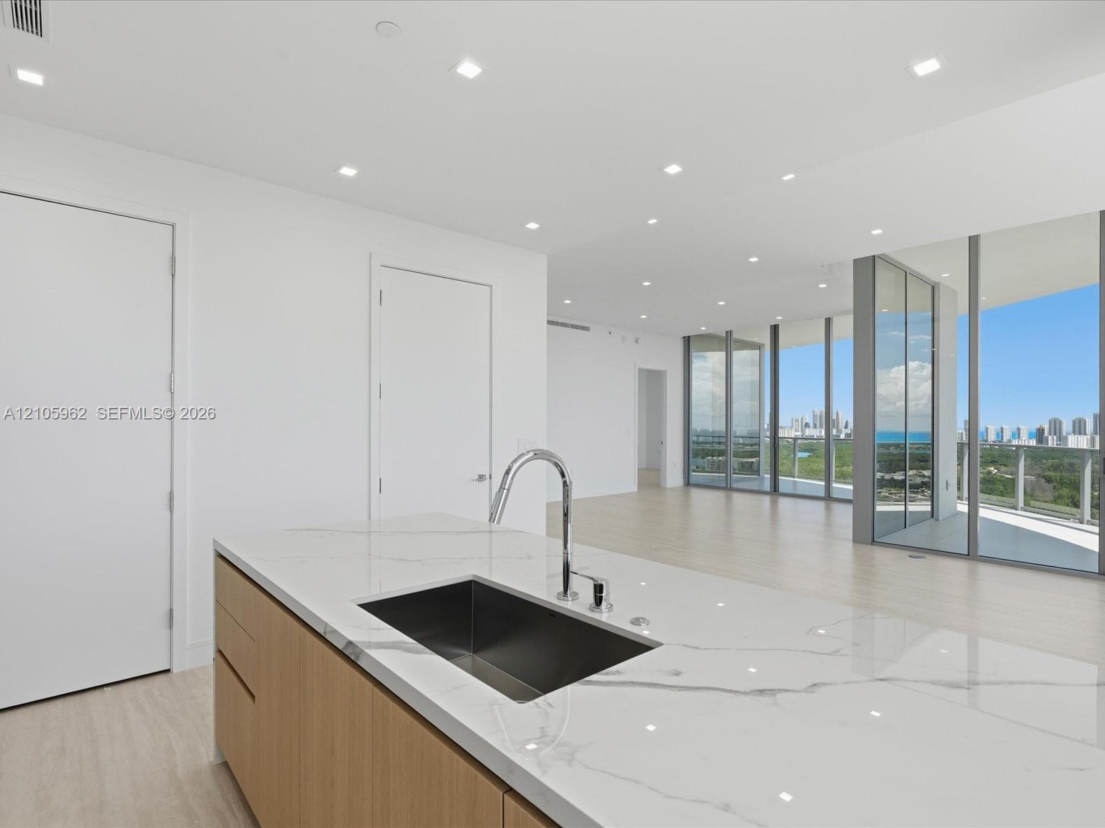 Kitchen island featuring a waterfall-edge stone countertop, undermount stainless steel sink, chrome pull-down faucet, light wood-finish cabinetry, and recessed lighting