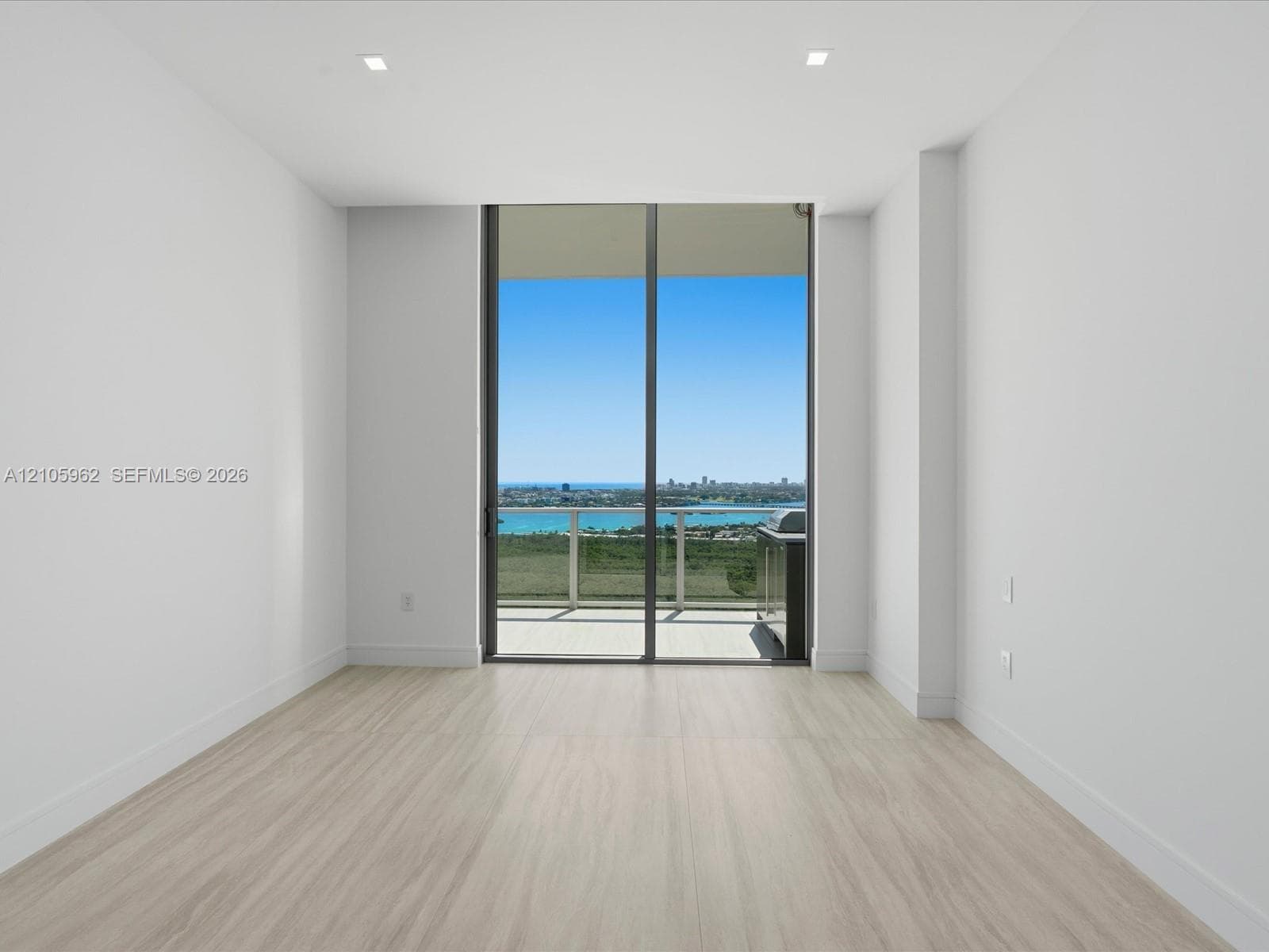 Floor-to-ceiling sliding glass doors, light-toned wood-finish flooring, recessed lighting, and balcony access with water and city skyline scenery