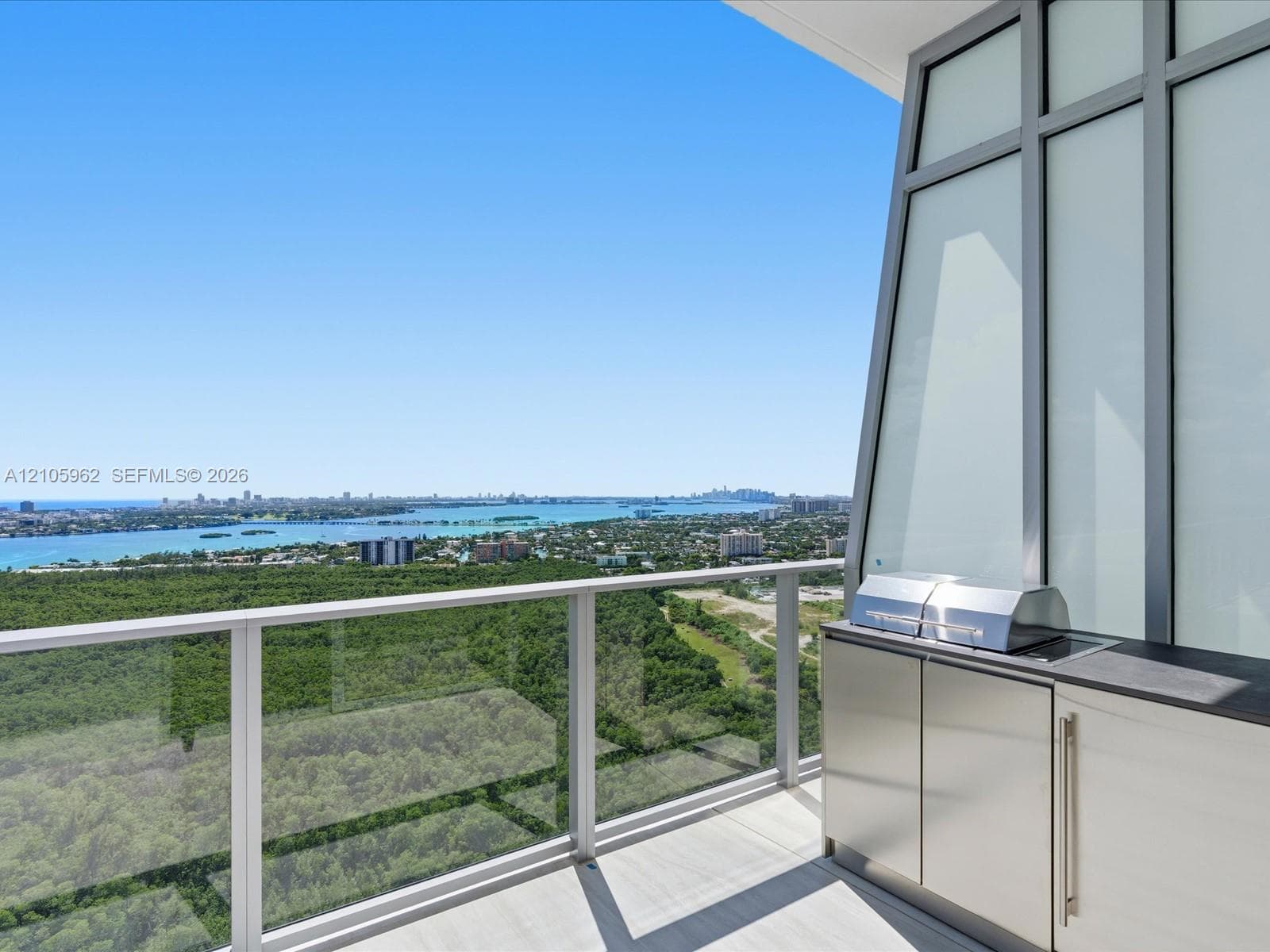 Covered terrace featuring glass panel railings, built-in outdoor kitchen with stainless steel grill, cabinetry with metal pulls, and stone-finish countertop