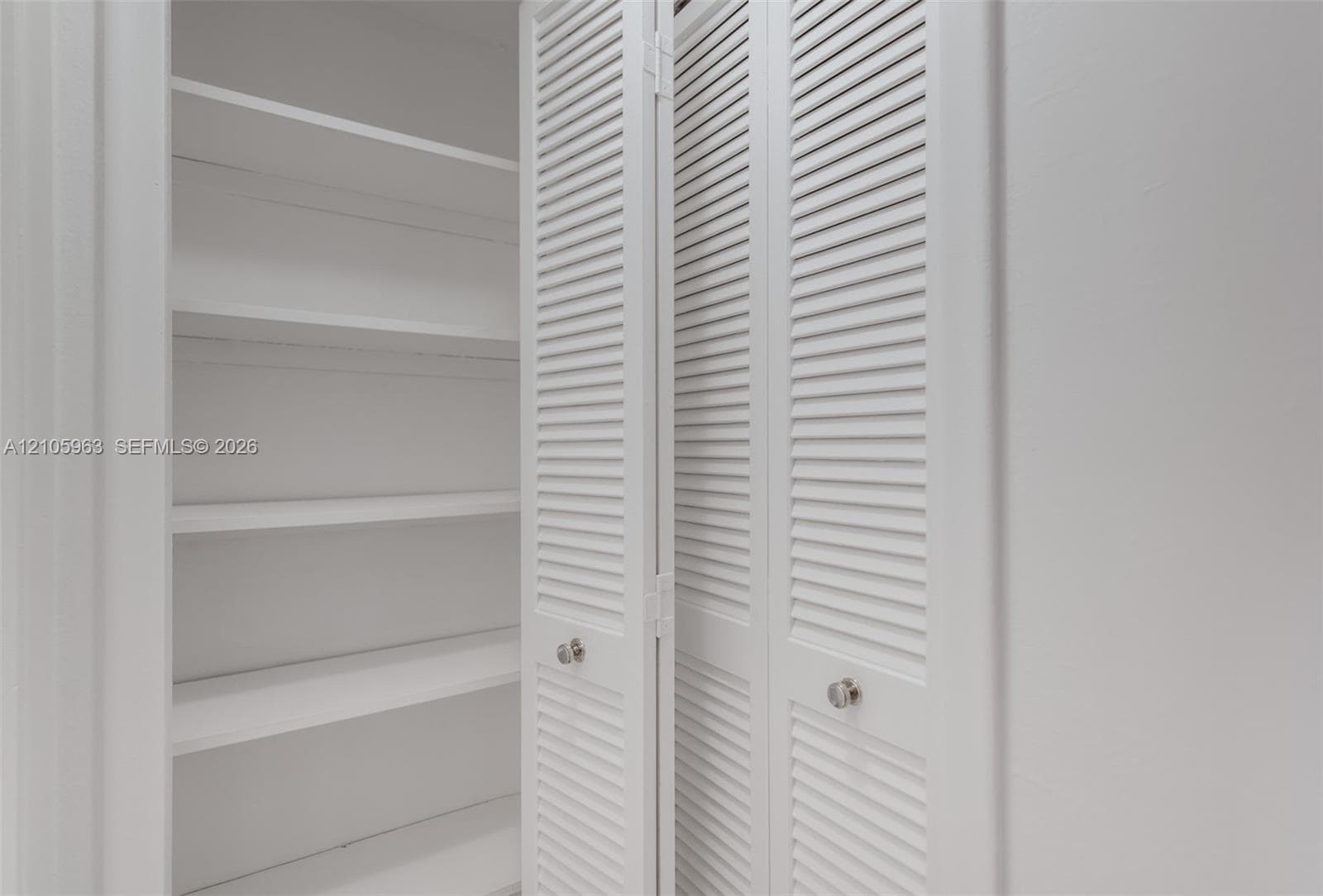 Storage closet featuring white louvered bifold doors, chrome round knobs, and interior shelving