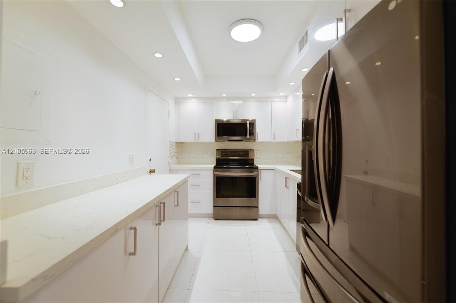 Galley kitchen featuring white cabinetry, light-toned stone-finish countertops, stainless steel appliances, recessed ceiling lighting, and large-format tile flooring