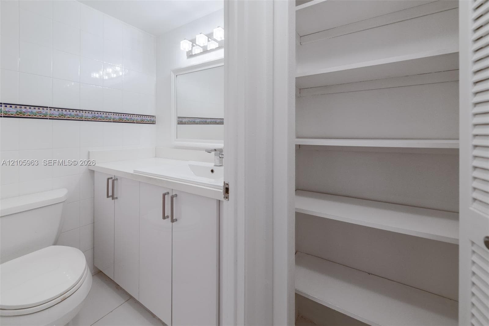 Bathroom featuring white cabinetry, integrated sink, wall-mounted mirror, ceramic tile walls with decorative border, and built-in shelving with louvered doors