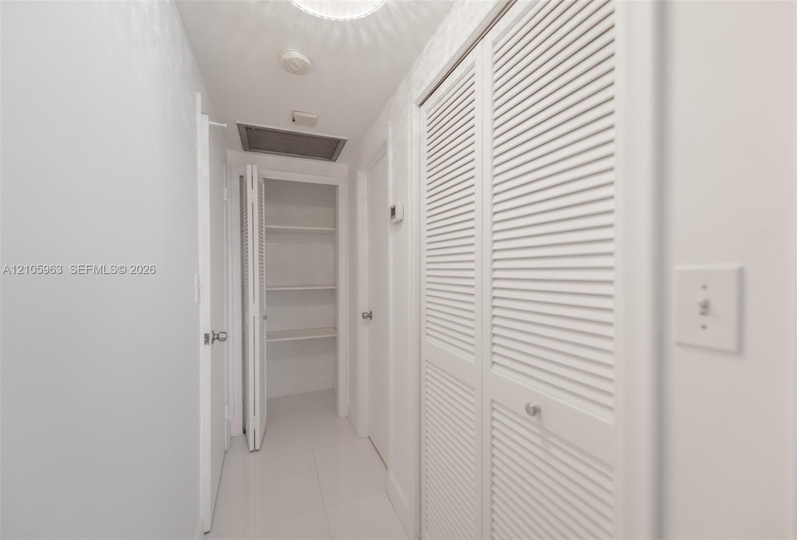 Hallway featuring white tile flooring, ceiling-mounted light fixture, and multiple sets of white louvered doors providing built-in storage