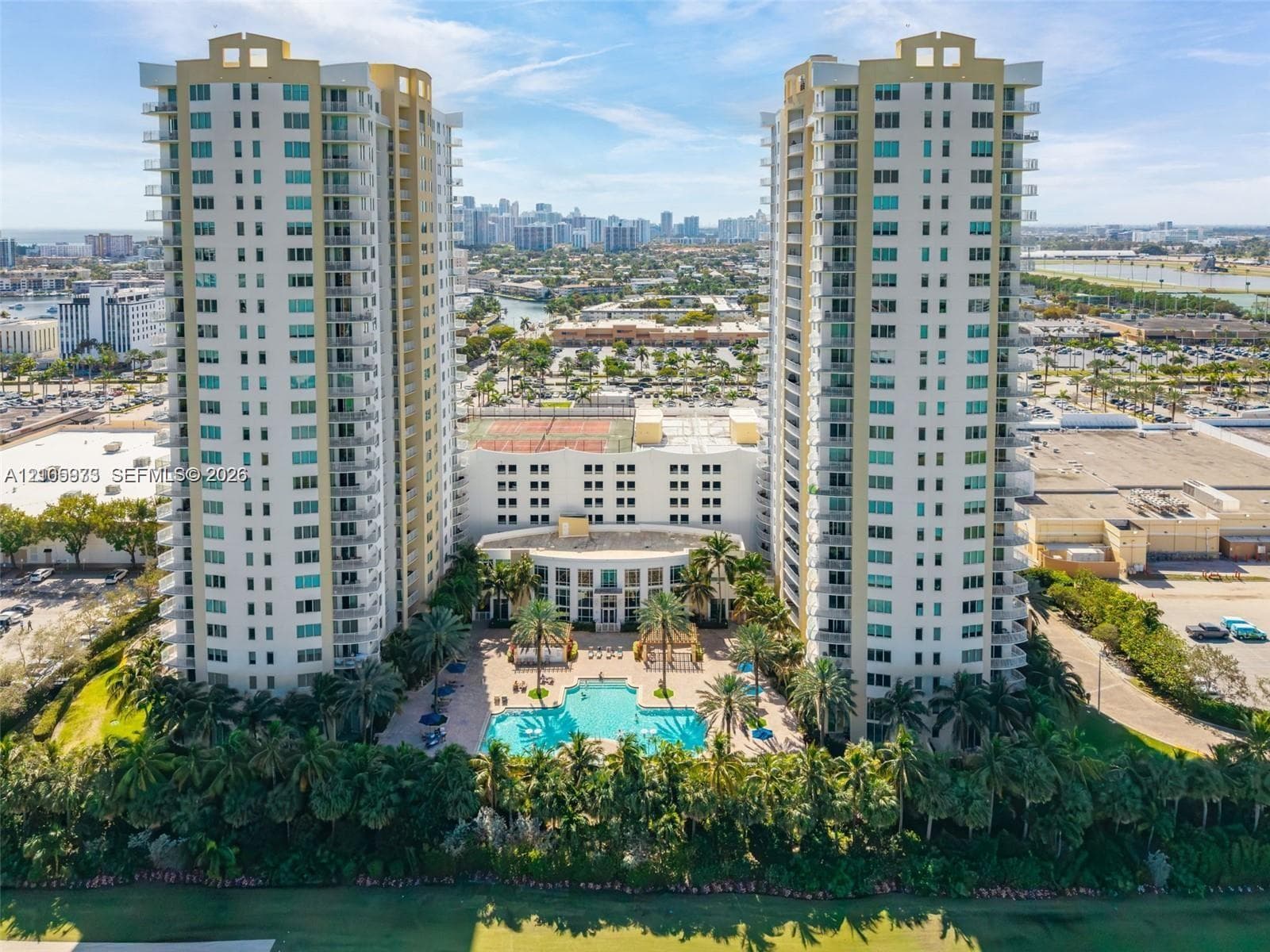 High-rise residential towers featuring multiple wrap-around balconies, a ground-level swimming pool, hardscaped patio areas, and tennis courts
