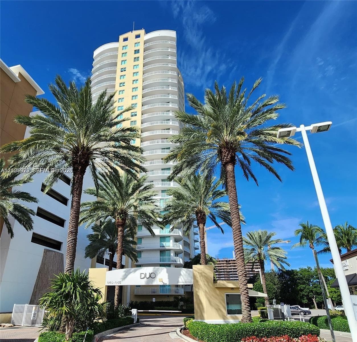 High-rise residential tower featuring white structural balconies, a neutral-toned facade, gated entrance with security kiosk, and mature palm tree landscaping