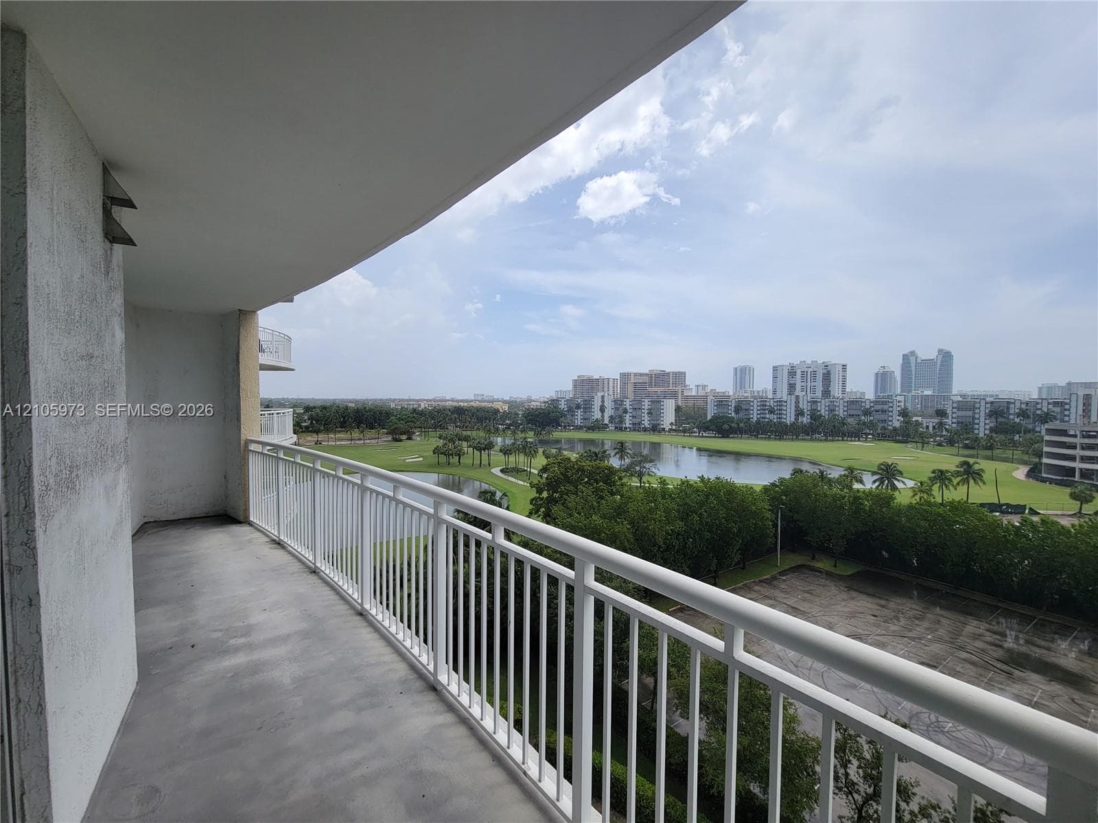 Covered balcony featuring concrete flooring and a white metal railing, overlooking a landscaped water feature and surrounding cityscape