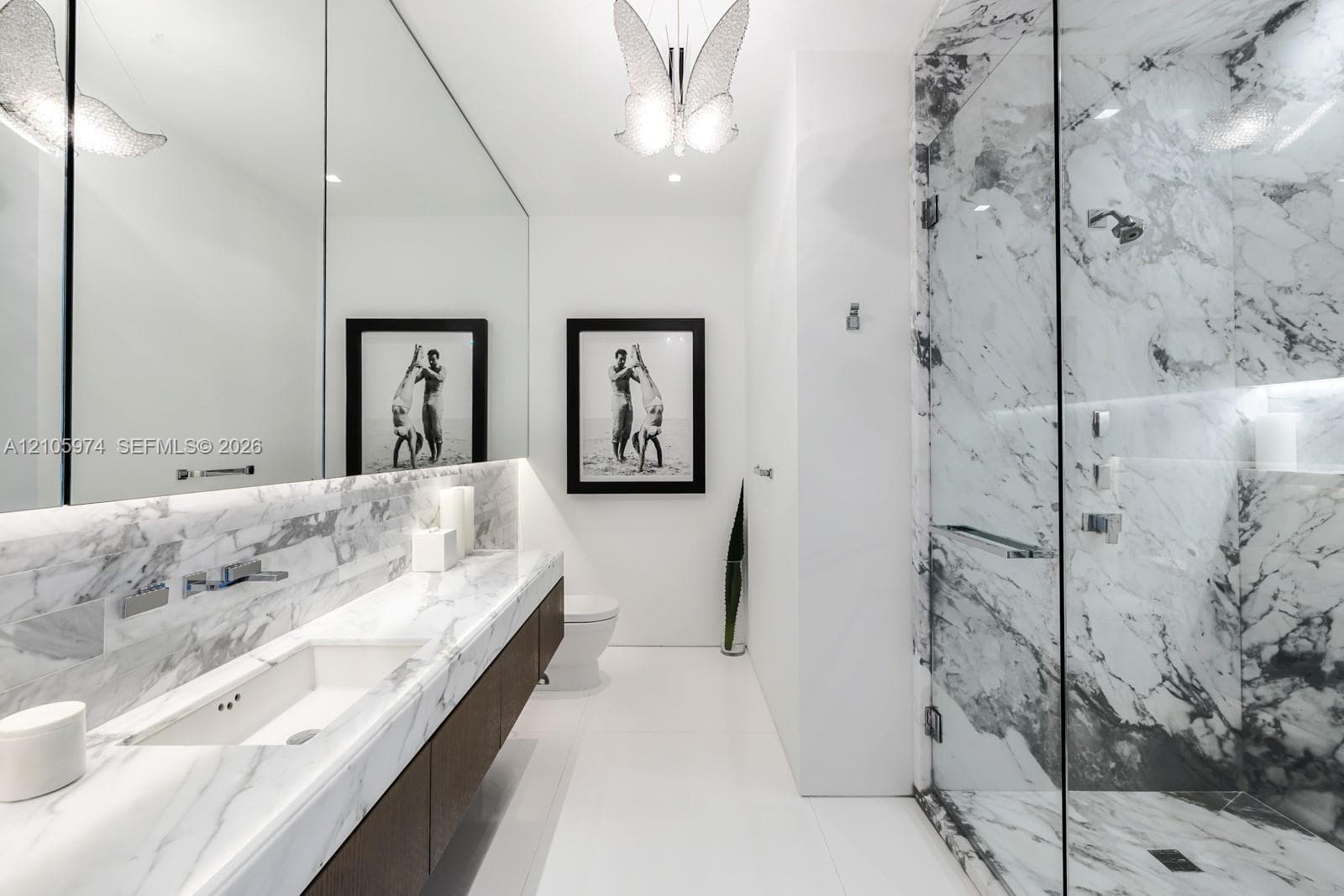 Stone-surfaced vanity with an integrated basin, wall-mounted faucet, expansive wall-to-wall mirror, walk-in shower with glass enclosure, and decorative overhead lighting fixture