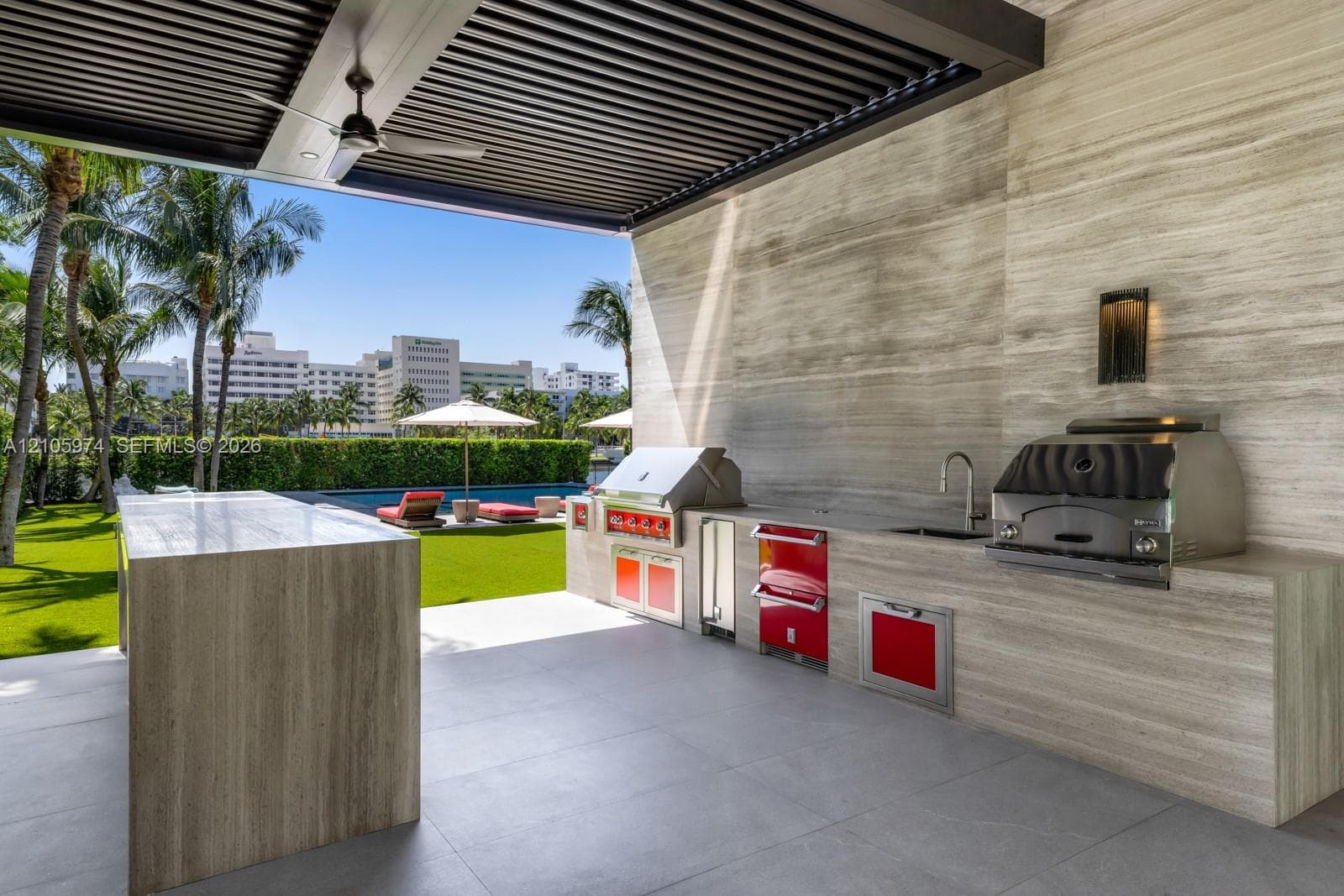 Covered outdoor kitchen featuring stone-clad surfaces, stainless steel built-in grill and beverage refrigeration, integrated faucet, and an overhead louvered pergola with a ceiling fan