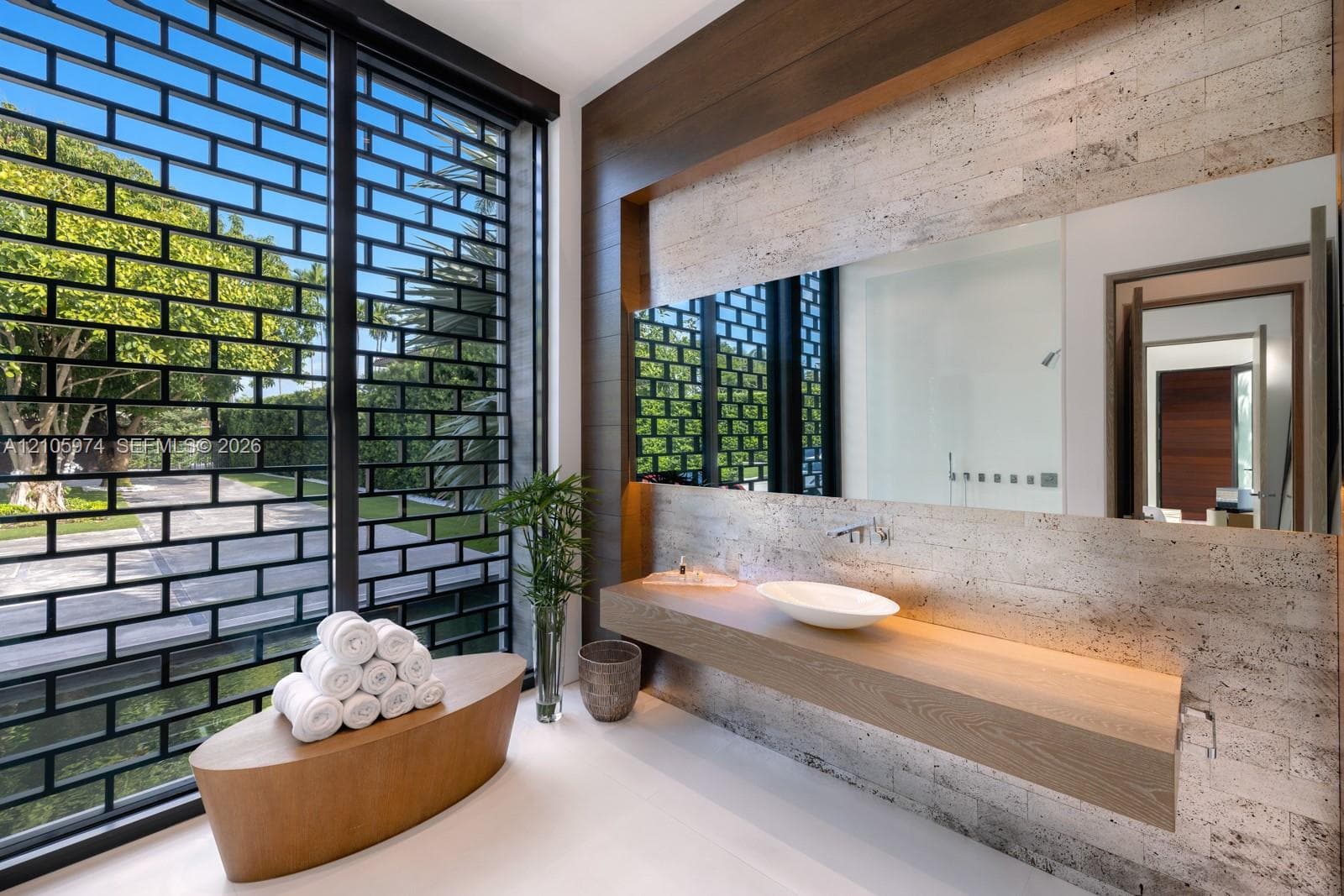 Floating wood-finish vanity with a white vessel sink, wall-mounted faucet, oversized frameless mirror, textured stone-tiled wall, and full-height exterior window with integrated metal grid screen